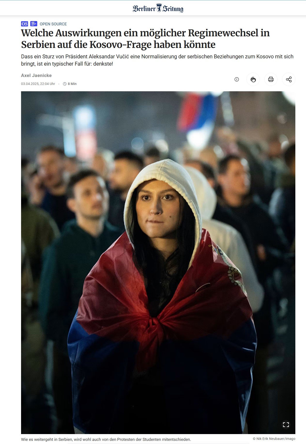 A woman in a hoodie and a flag wrapped around her shoulders standing among a crowd of people during what appears to be a protest or rally at night.