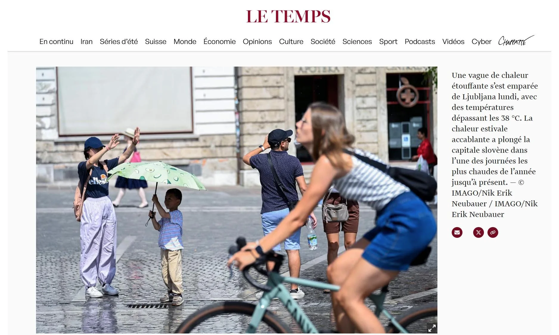 People walking and cycling on a city street on a hot day, with one woman holding a child with an umbrella, and others dressed in summer clothes.