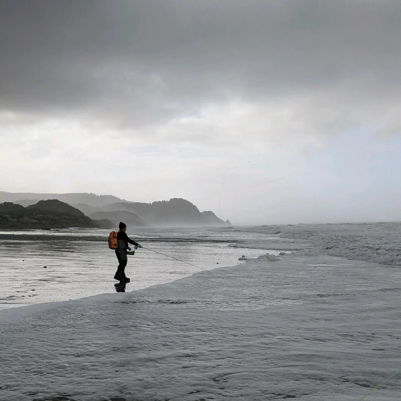 Person fishing on a foggy beach with a rocky coastline in the background.