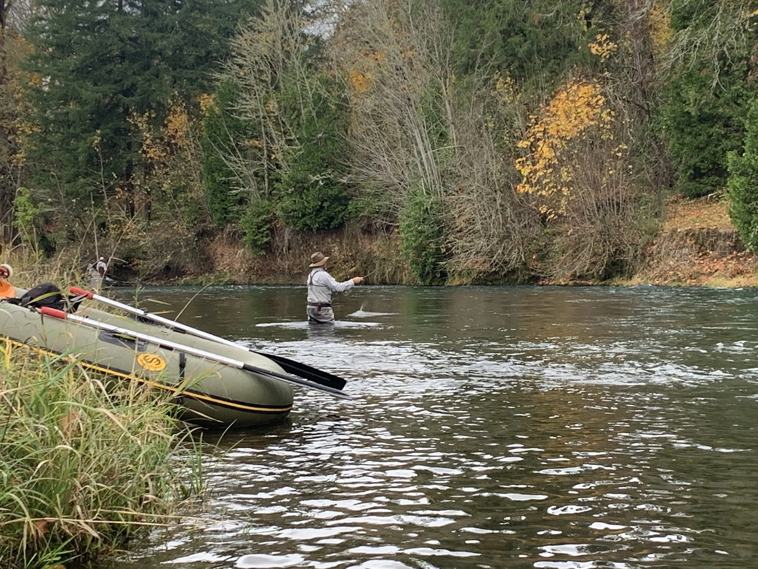 Person fly fishing in a river with inflatable raft nearby, surrounded by autumn trees.