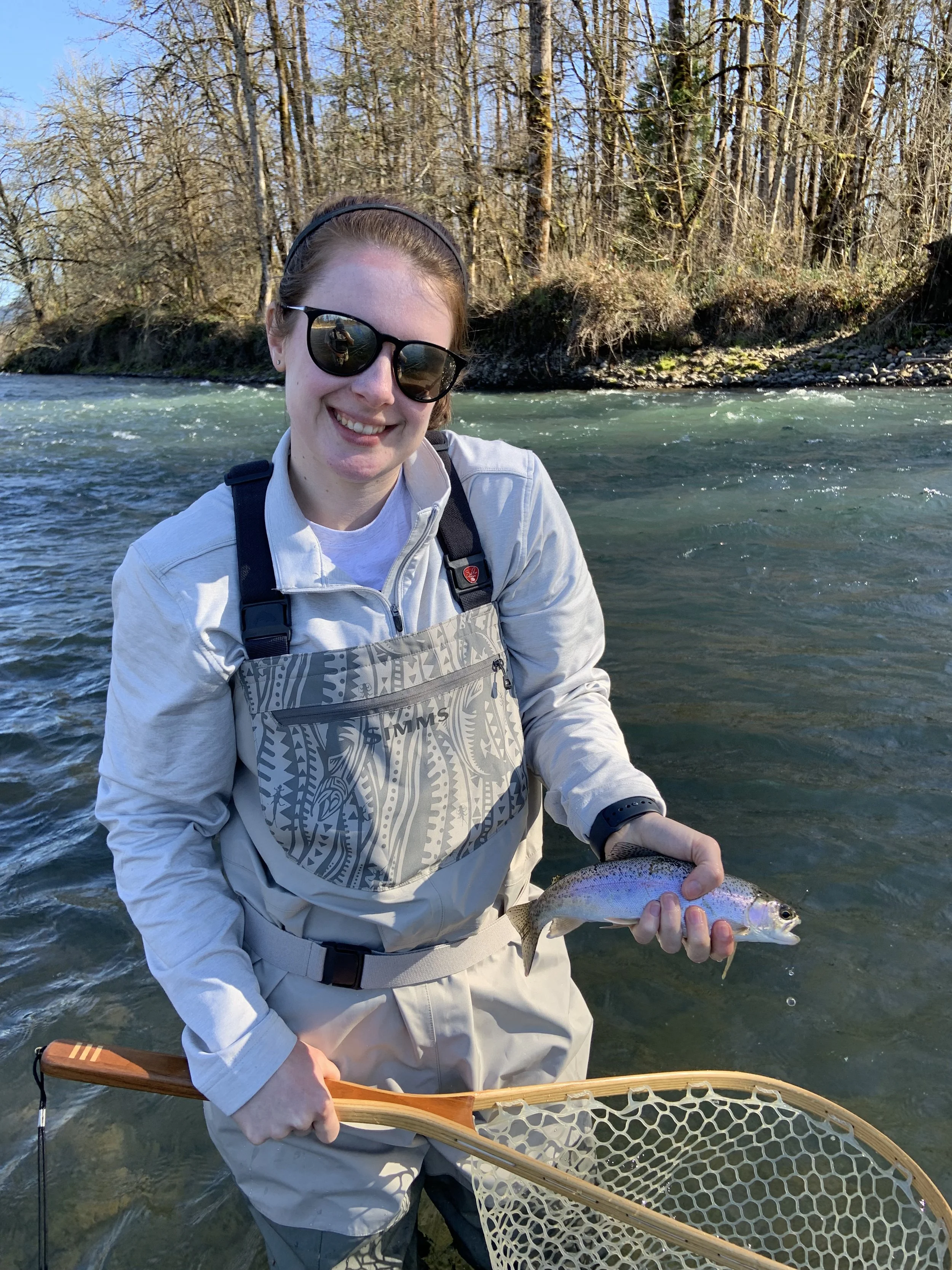 Person wearing sunglasses and waders holding a fish and fishing net by a river.