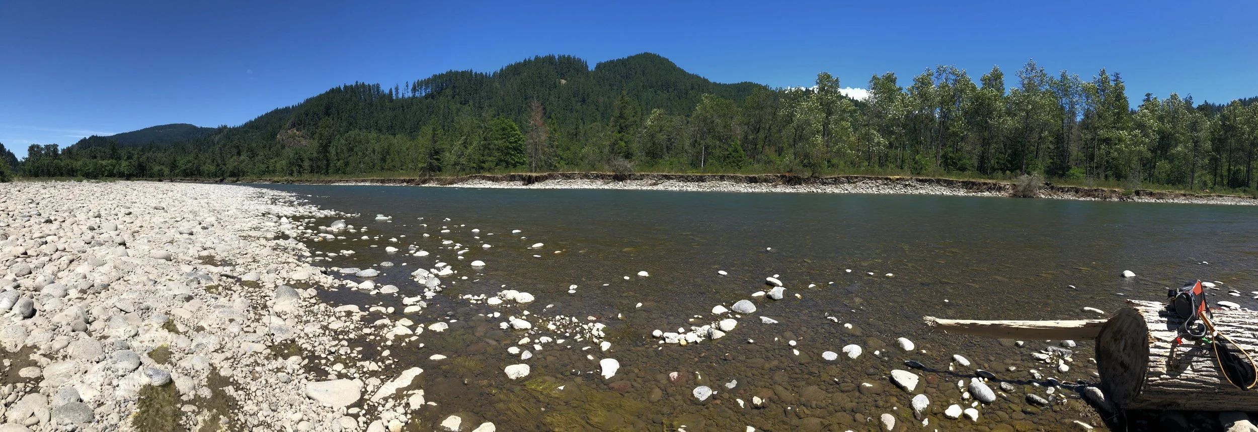Scenic view of a river with rocky bank and forested hills in the background under a clear blue sky.