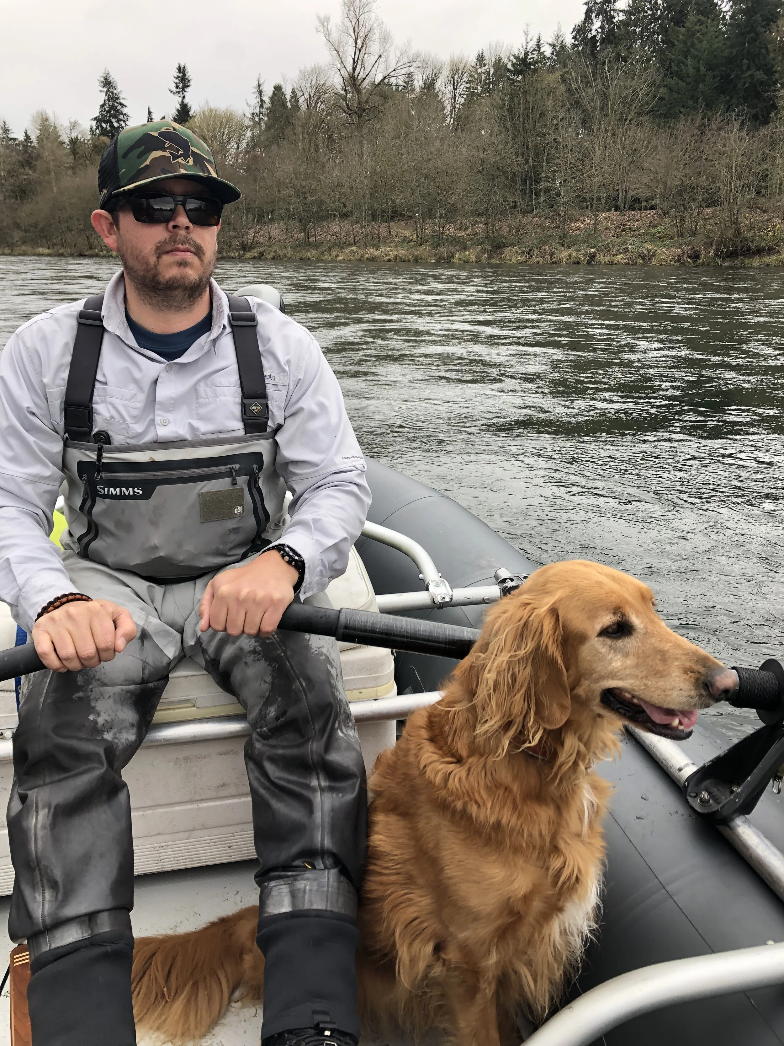Man in fishing gear with a golden retriever on a boat in a river.