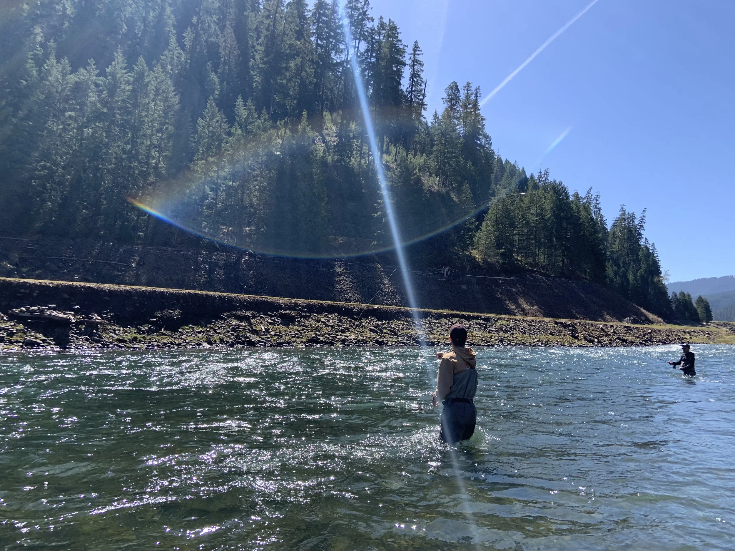 Two people fishing in a river under a bright sunbeam, surrounded by a forested hillside.