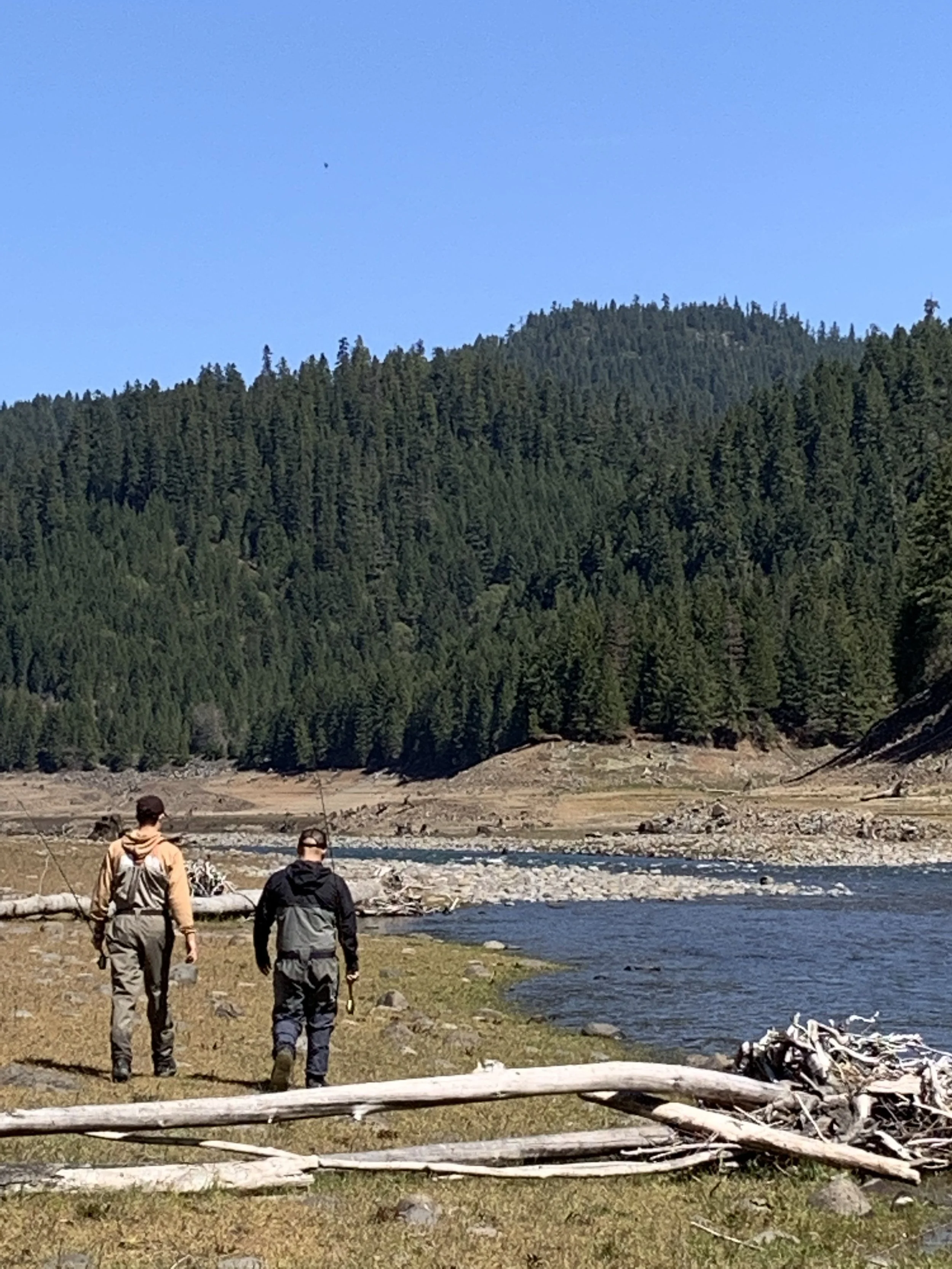 Two people walking along a riverbank with a forested hill in the background