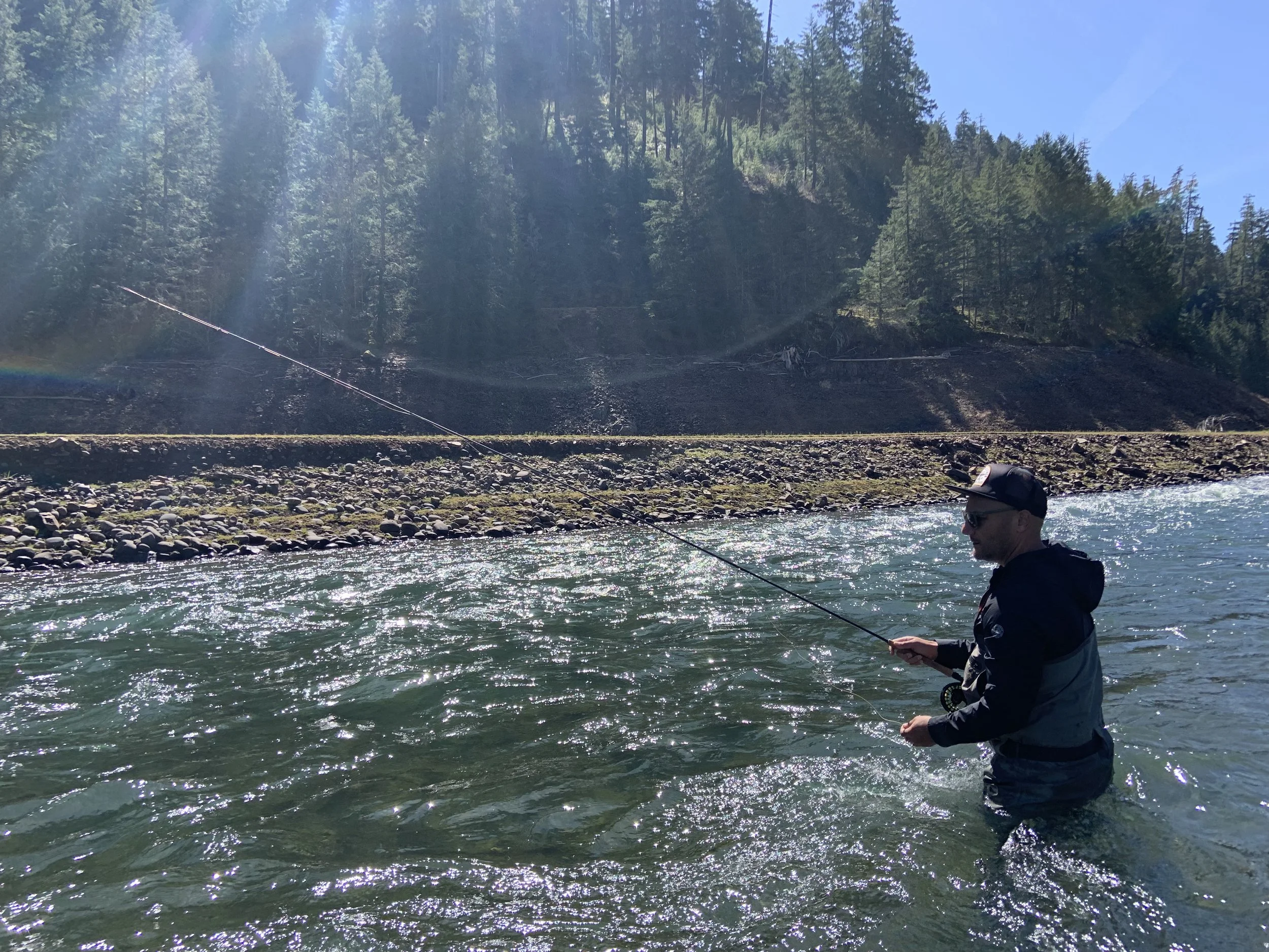 Man fly fishing in a river with rocky shore and forested background.