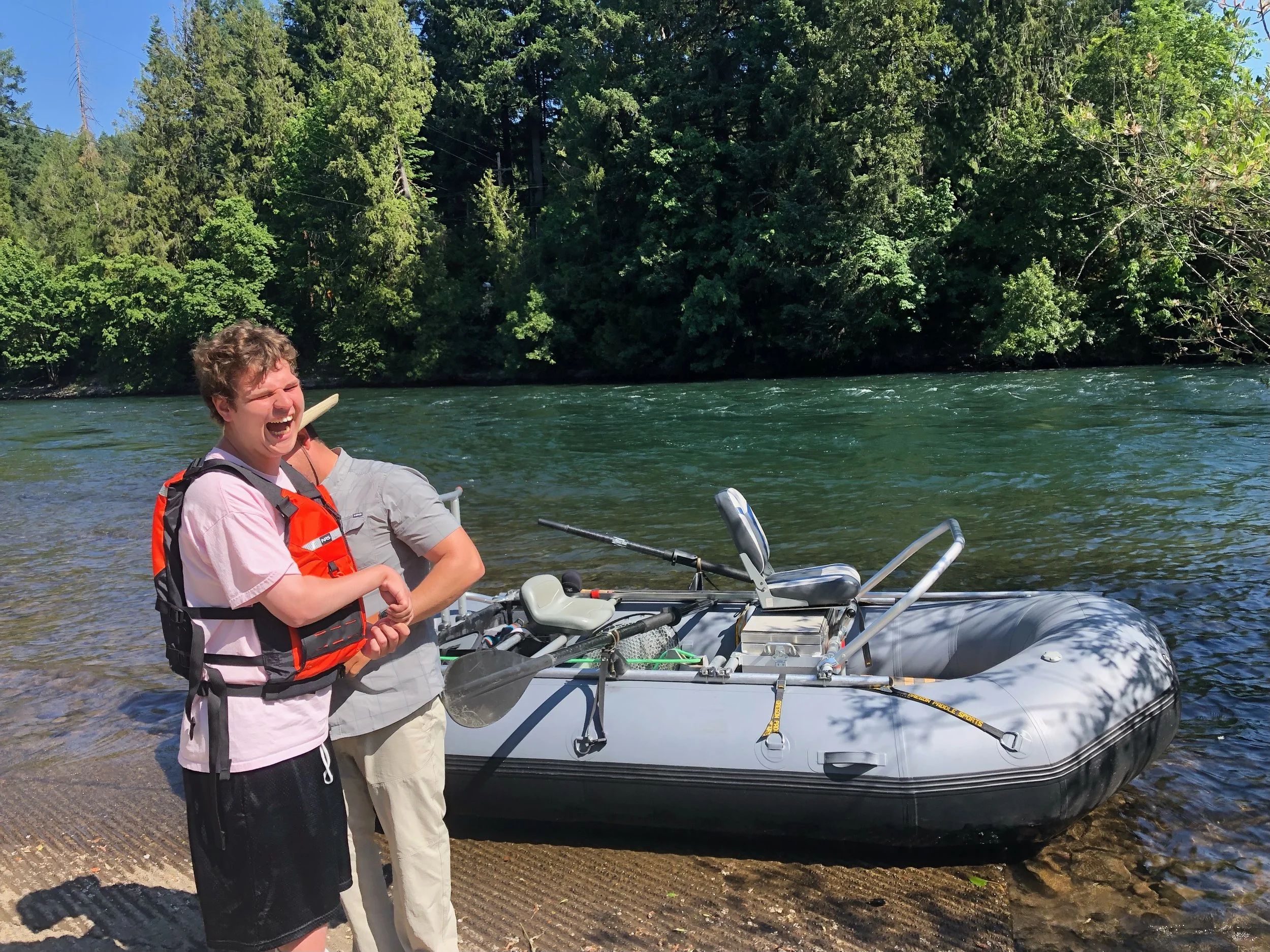 Two people standing near an inflatable boat by a river with trees in the background, one wearing an orange life vest.
