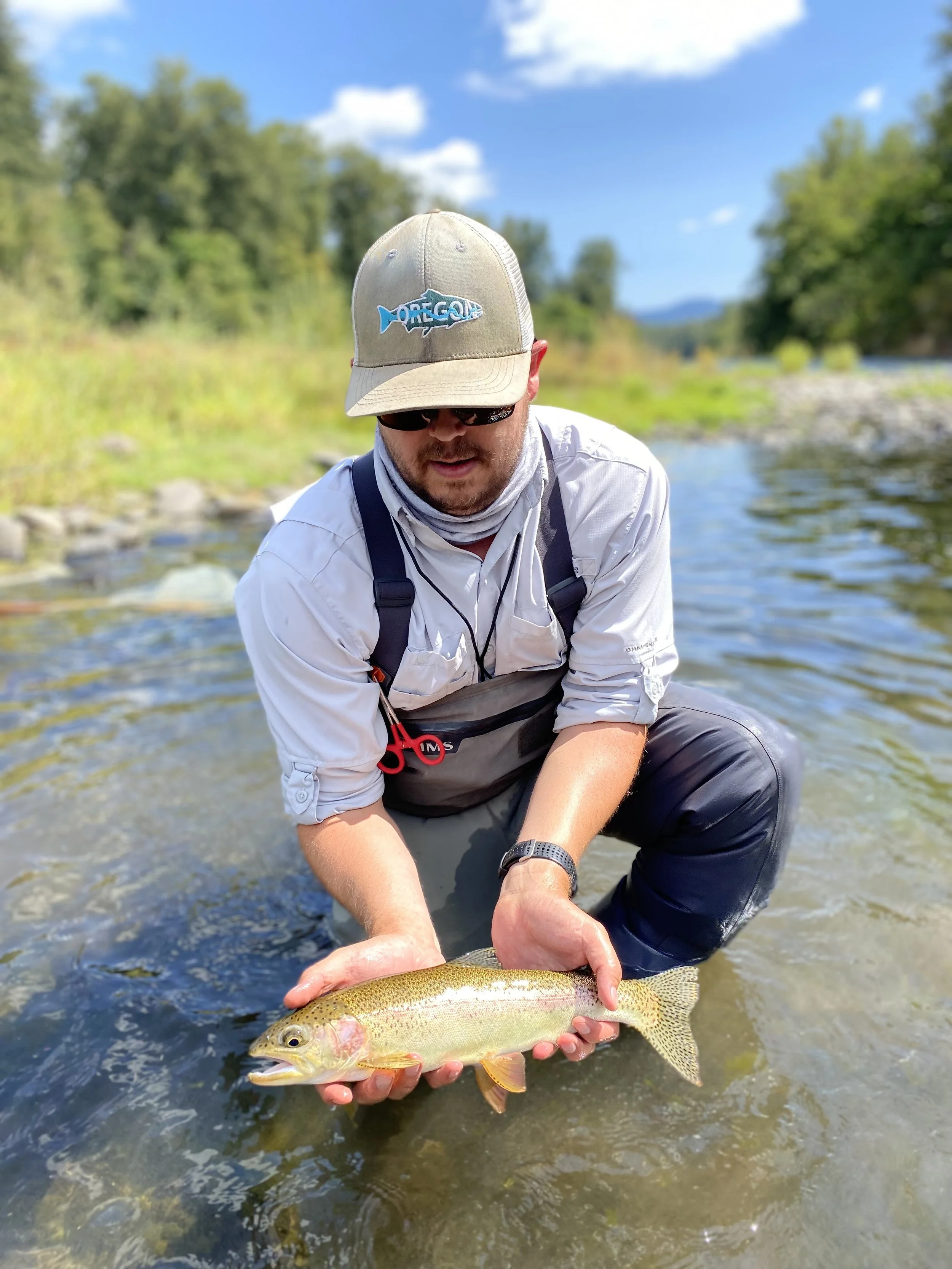 Person holding a fish while standing in a river, wearing a hat and fishing gear, with trees and blue sky in the background.