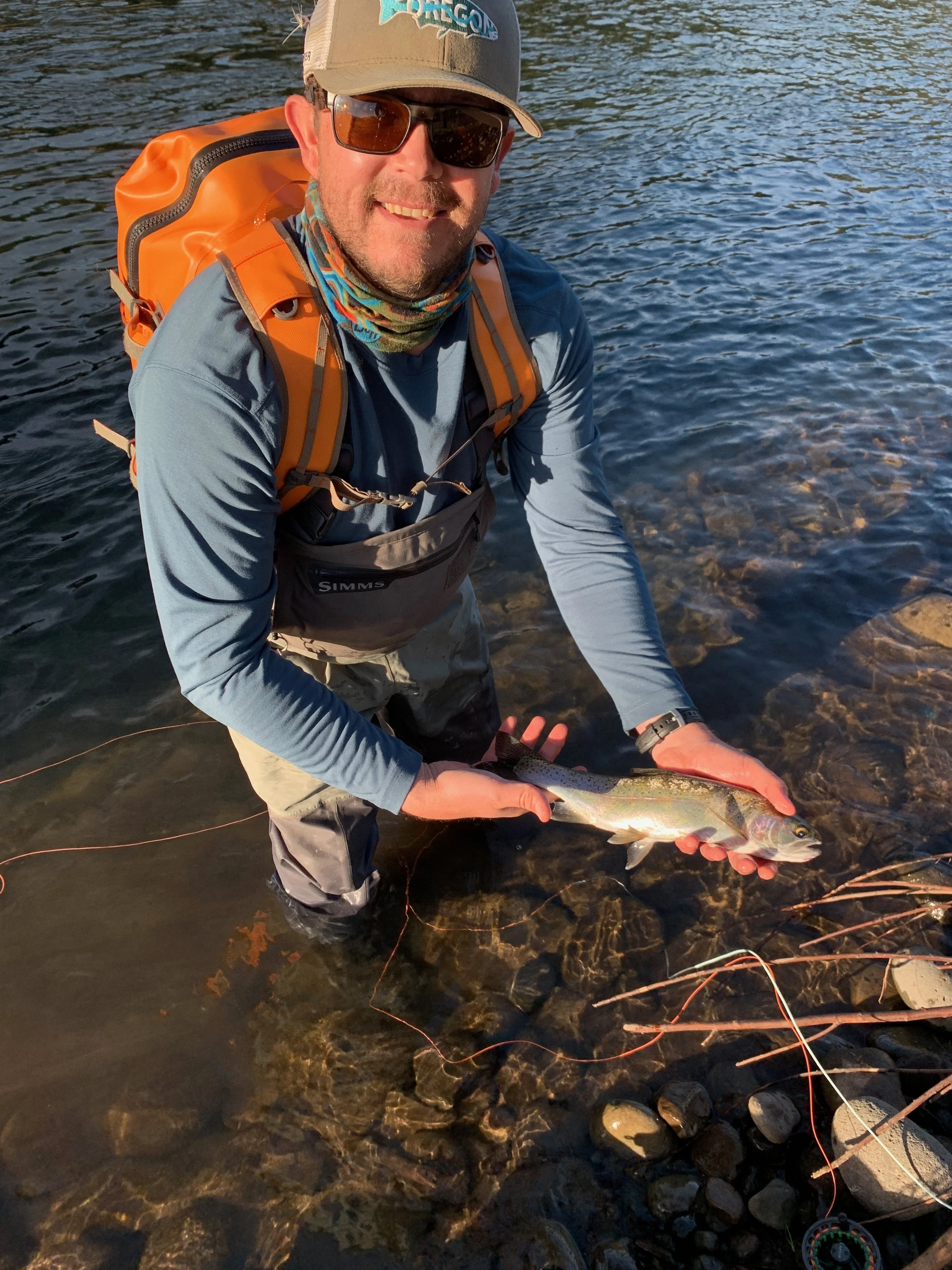 Man holding a fish standing in a river, wearing outdoor gear and an orange backpack.
