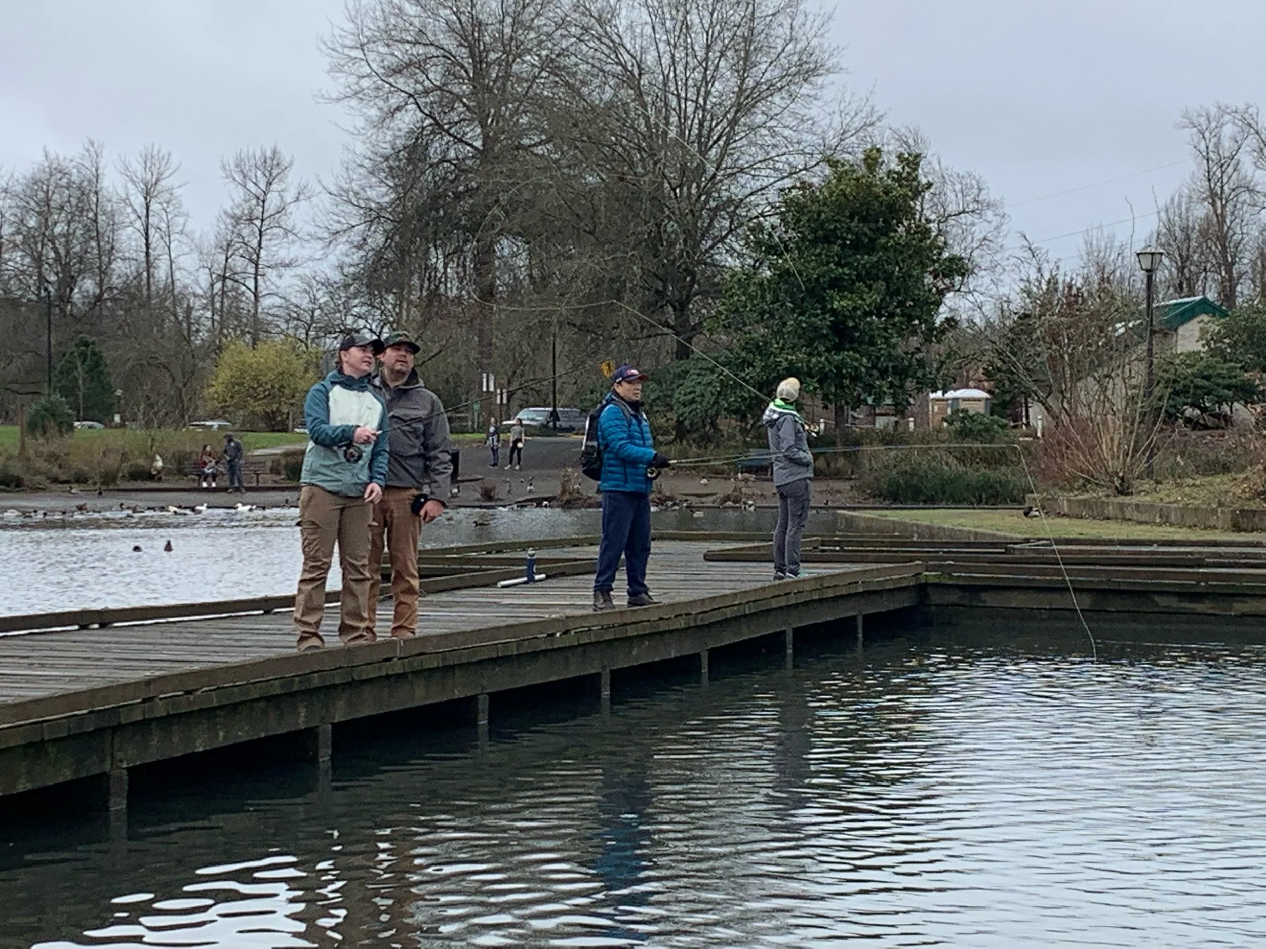People fishing on a wooden dock near a lake with trees in the background.