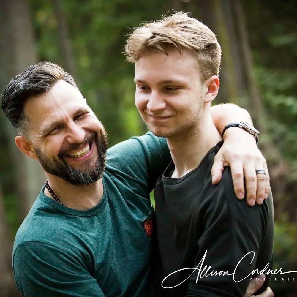 Craig Wanless and his son Jadon are smiling and hugging outdoors in a forest.