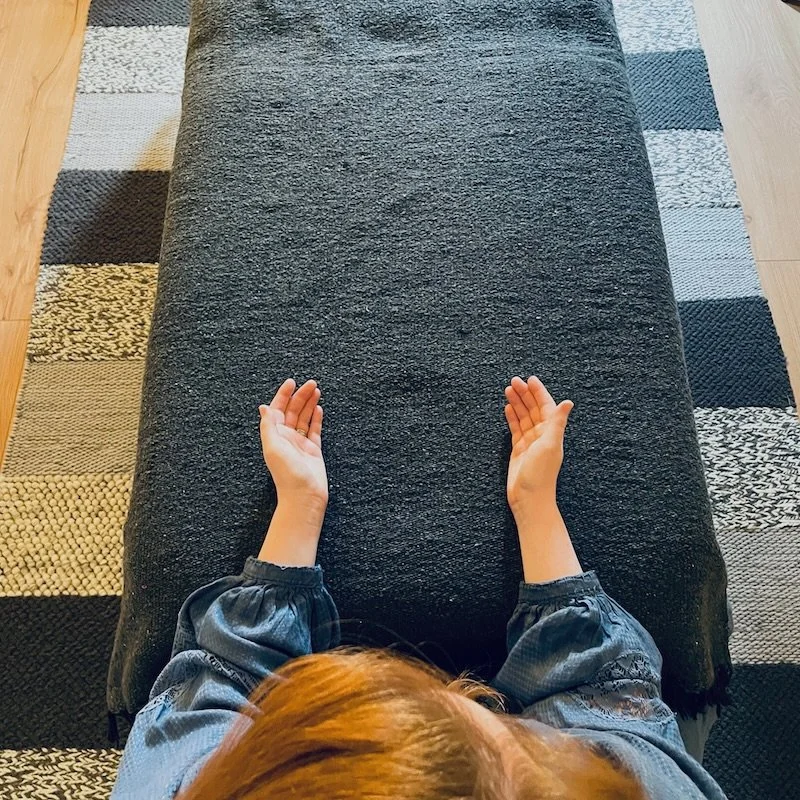 Overhead view of a person with red hair sitting at a black massage or therapy table, with hands resting on the table, in front of a patterned rug on a wooden floor.