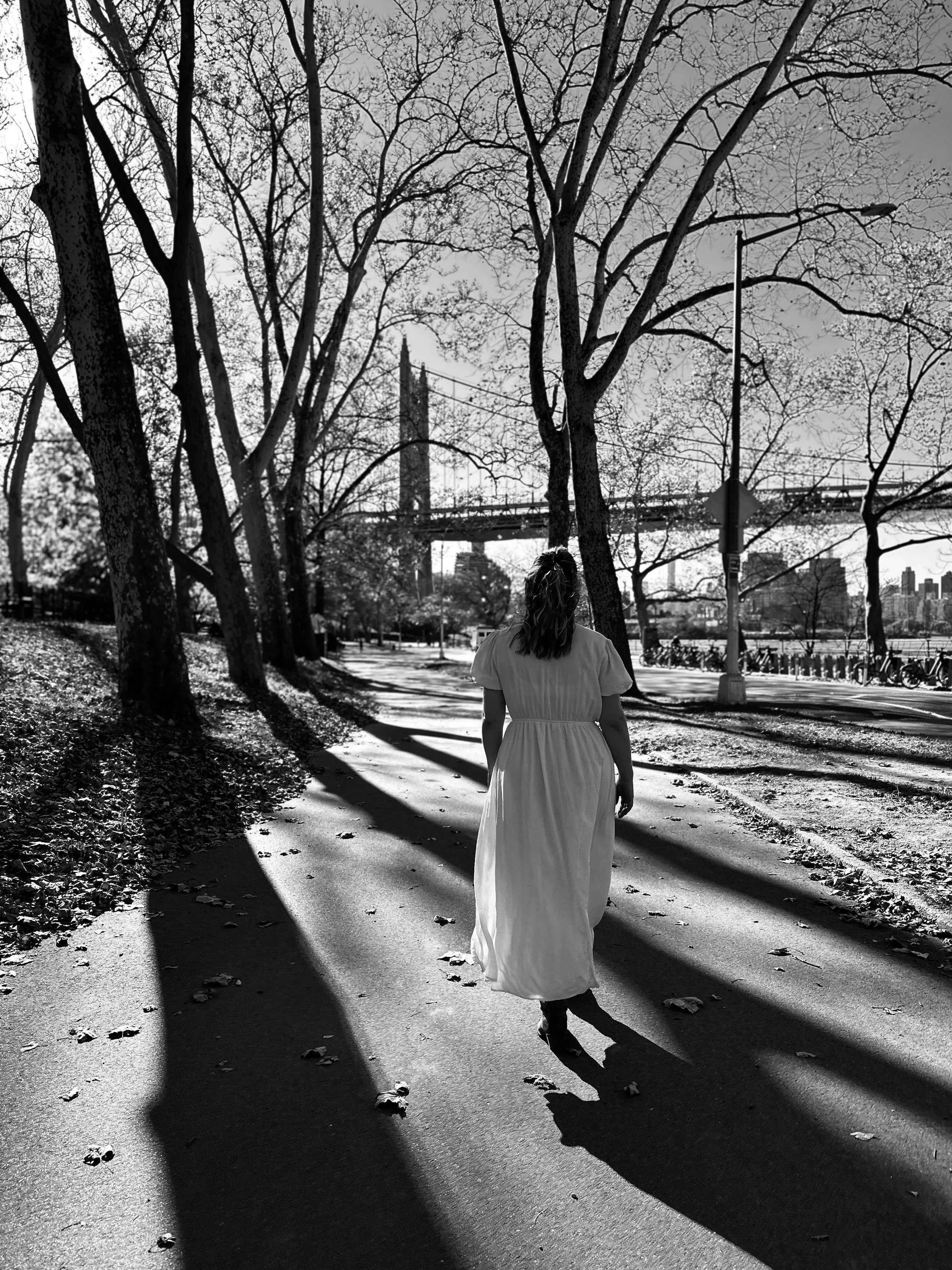 A woman walking on a sidewalk lined with trees, casting long shadows on the ground. In the background, a bridge and city skyline are visible.