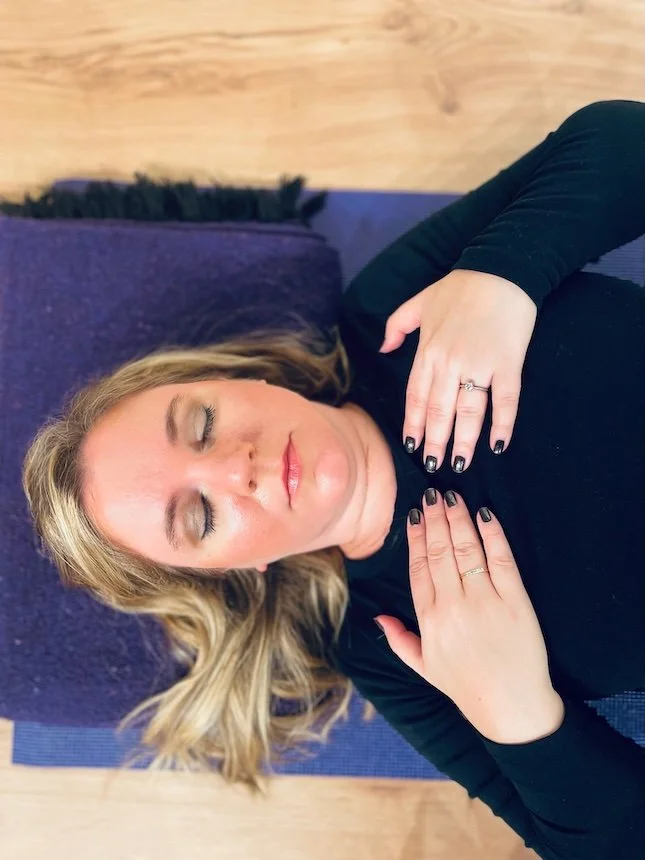 A woman with blonde hair, wearing black clothing, is lying down on a purple yoga mat with her eyes closed and hands resting on her chest, showing rings on her fingers.
