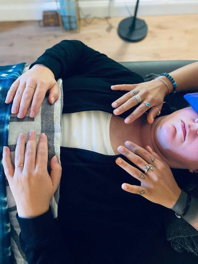 Close-up of a person lying on a couch with multiple hands placed on their chest and torso, wearing rings, bracelets, and a watch, with a background of wooden flooring and a standing lamp.
