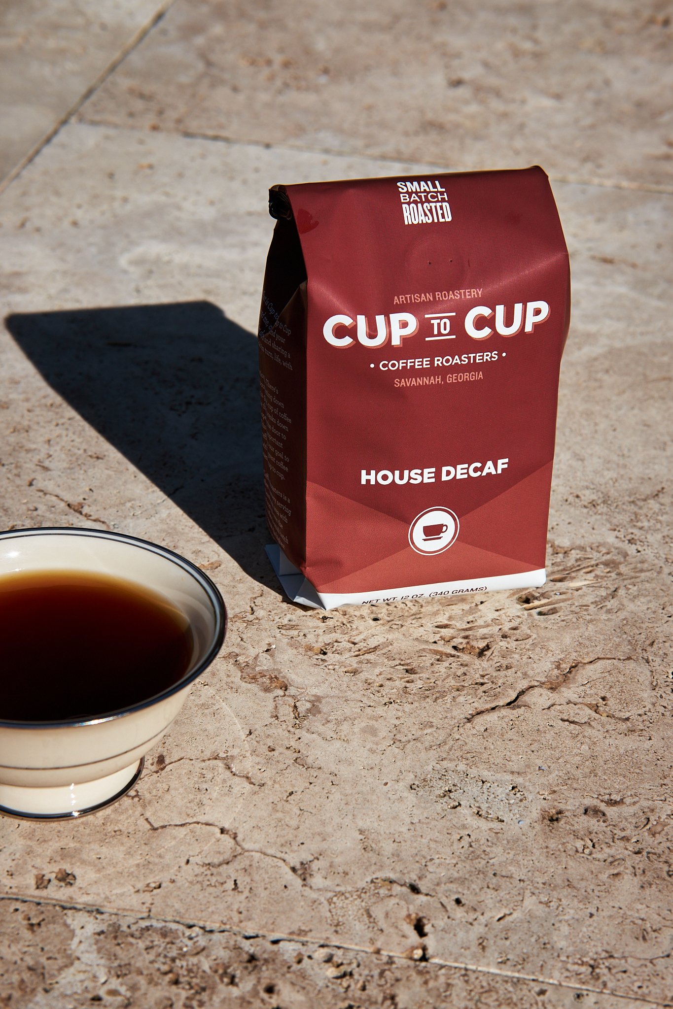 A cup of black coffee next to a bag of small batch roasted house decaf coffee from CUP TO CUP Coffee Roasters in Savannah, Georgia, on a textured outdoor surface.