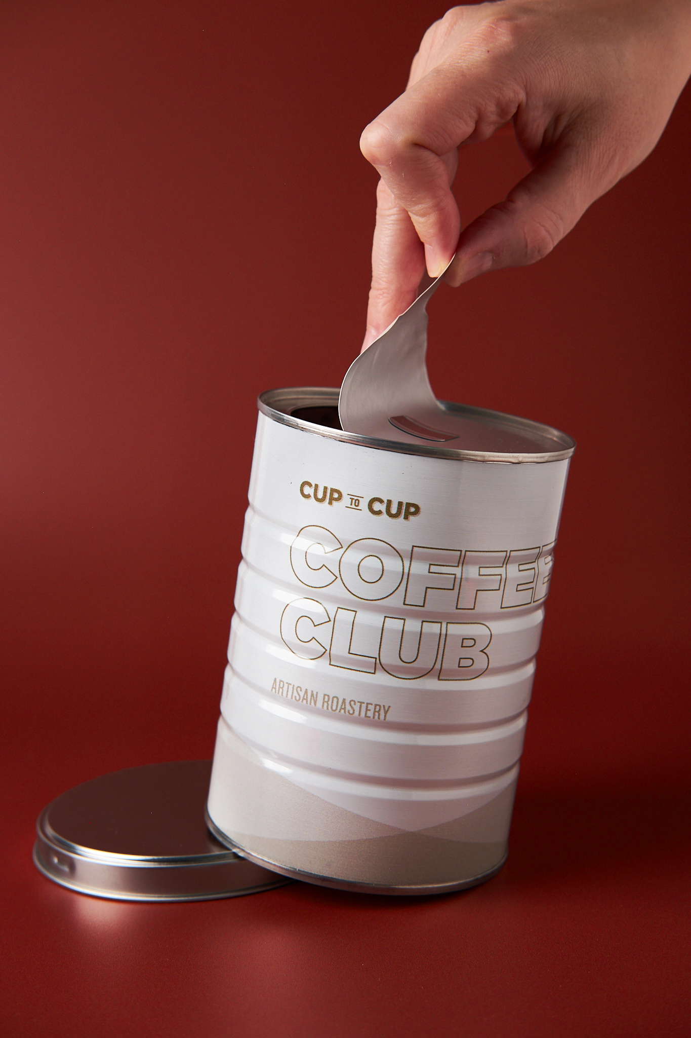 A hand opening a can of coffee labeled 'Coffee Club Artisan Roastery' against a red background.