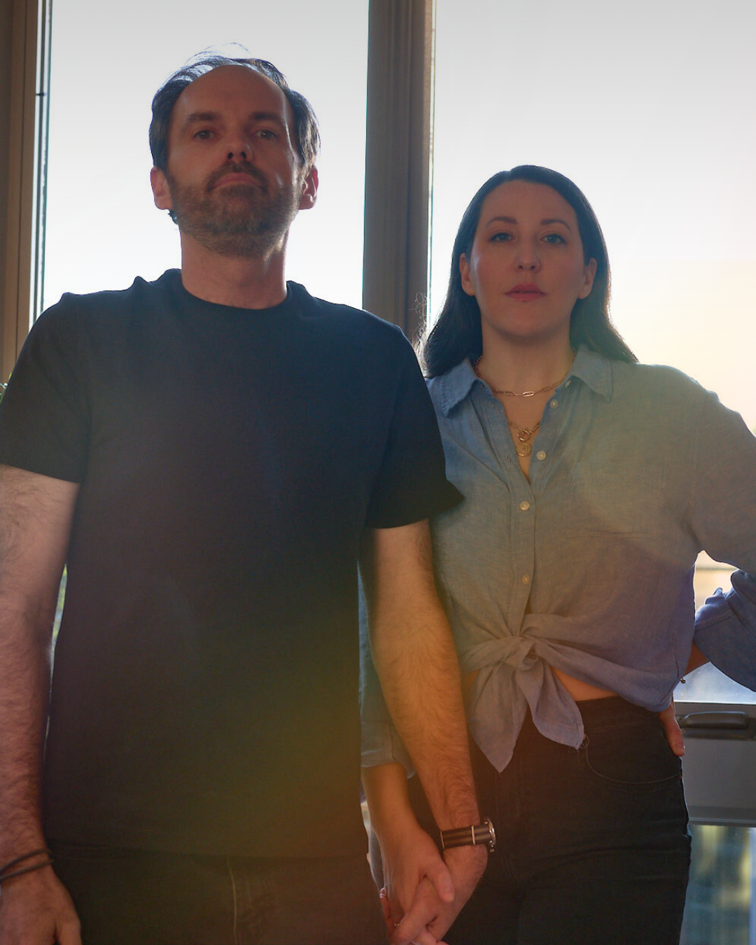A man and a woman holding hands indoors near a window with natural light, both looking serious.