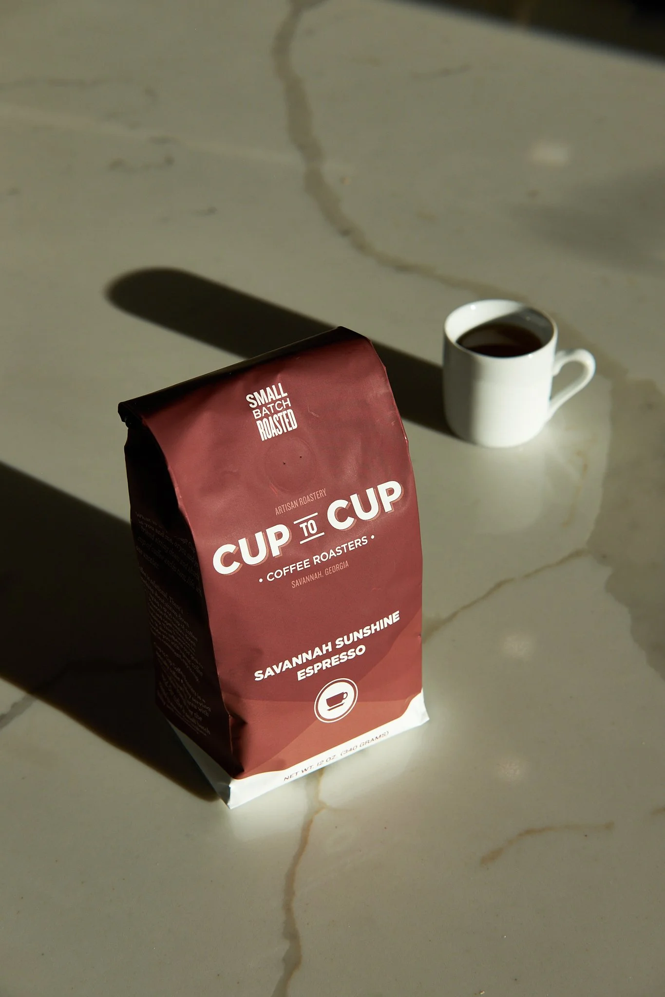 A bag of small batch roasted coffee labeled 'Cup to Cup, Savannah Sunshine Espresso,' placed on a light-colored marble surface, with a white mug filled with coffee in the background casting a shadow.