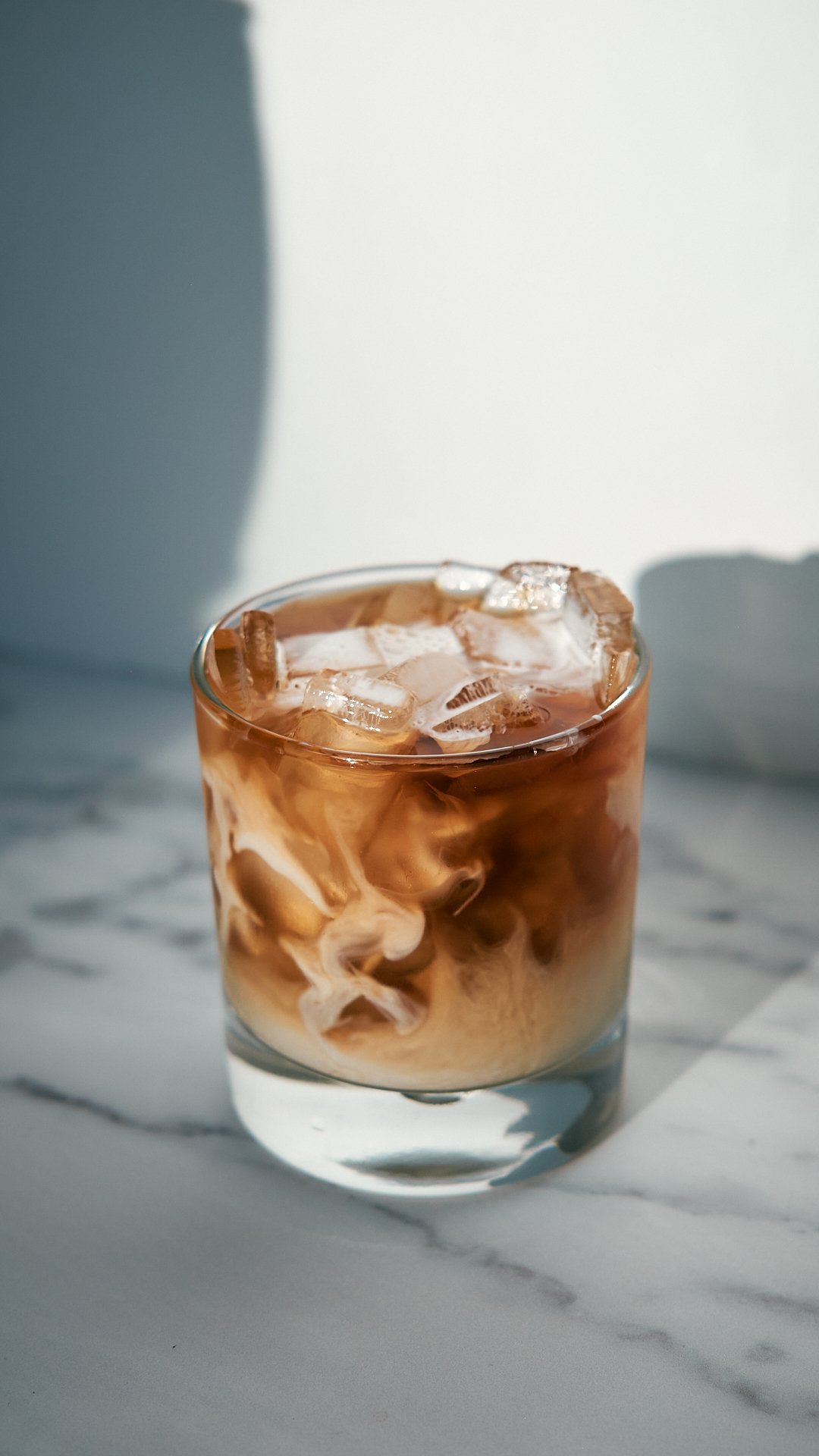 Glass of iced coffee with cream on a marble surface, natural light creating a shadow.
