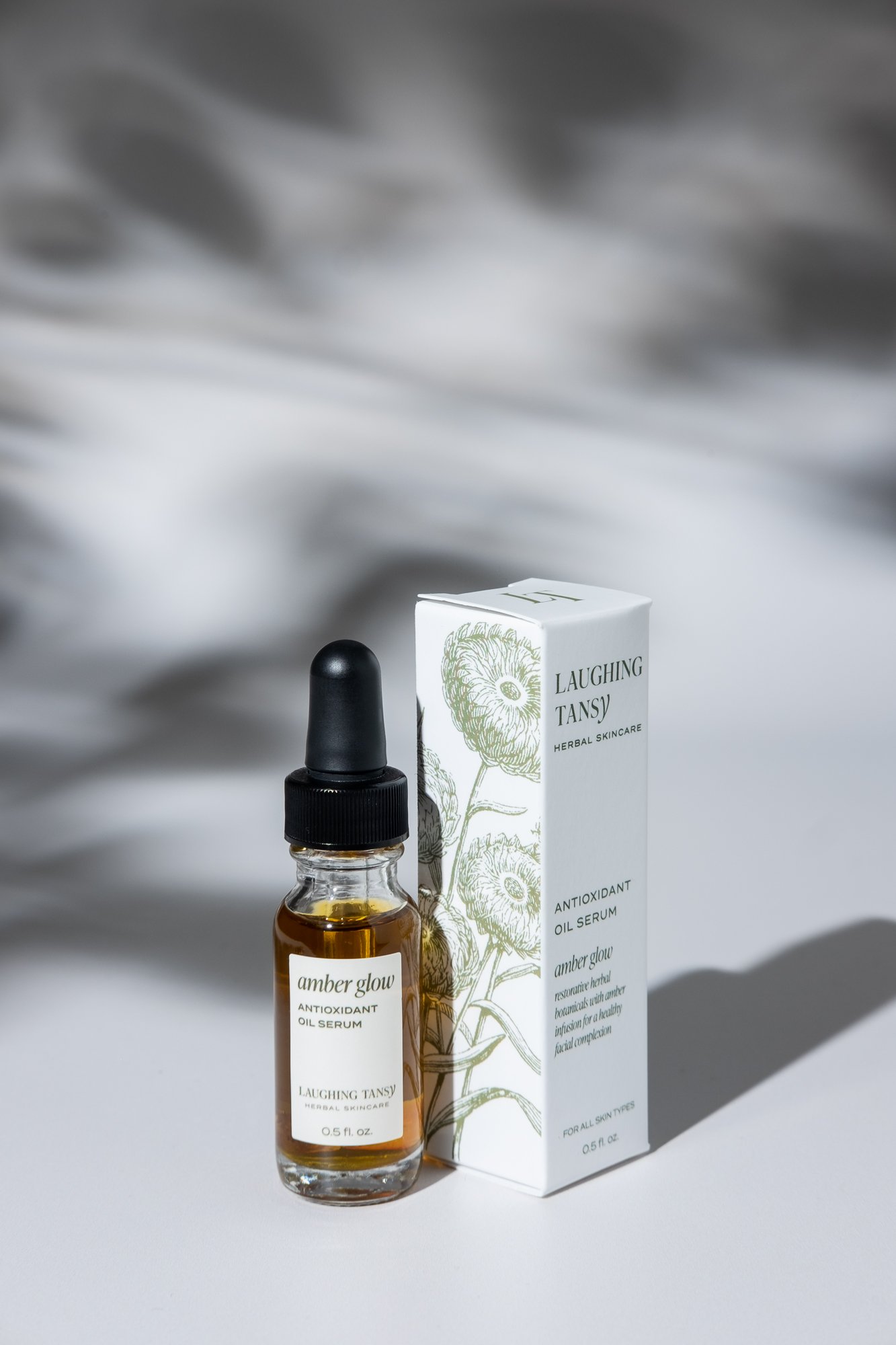 A small glass bottle of amber-colored antioxidant oil serum with a black dropper cap, alongside a white box featuring floral artwork and product details for Laughing Tansy herbal skincare.