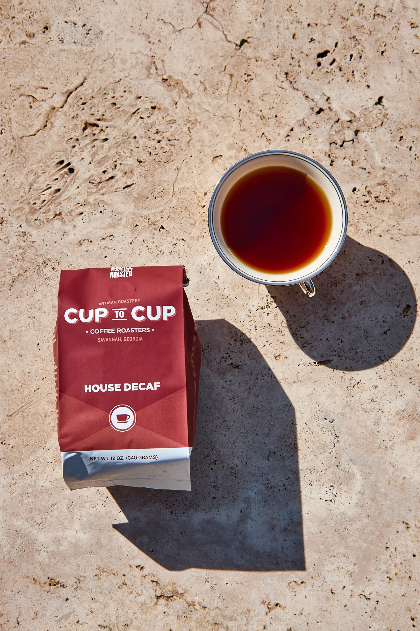 A cup of black coffee and a bag of Cup to Cup house decaf coffee on a sandy surface, with shadows cast by the coffee cup.