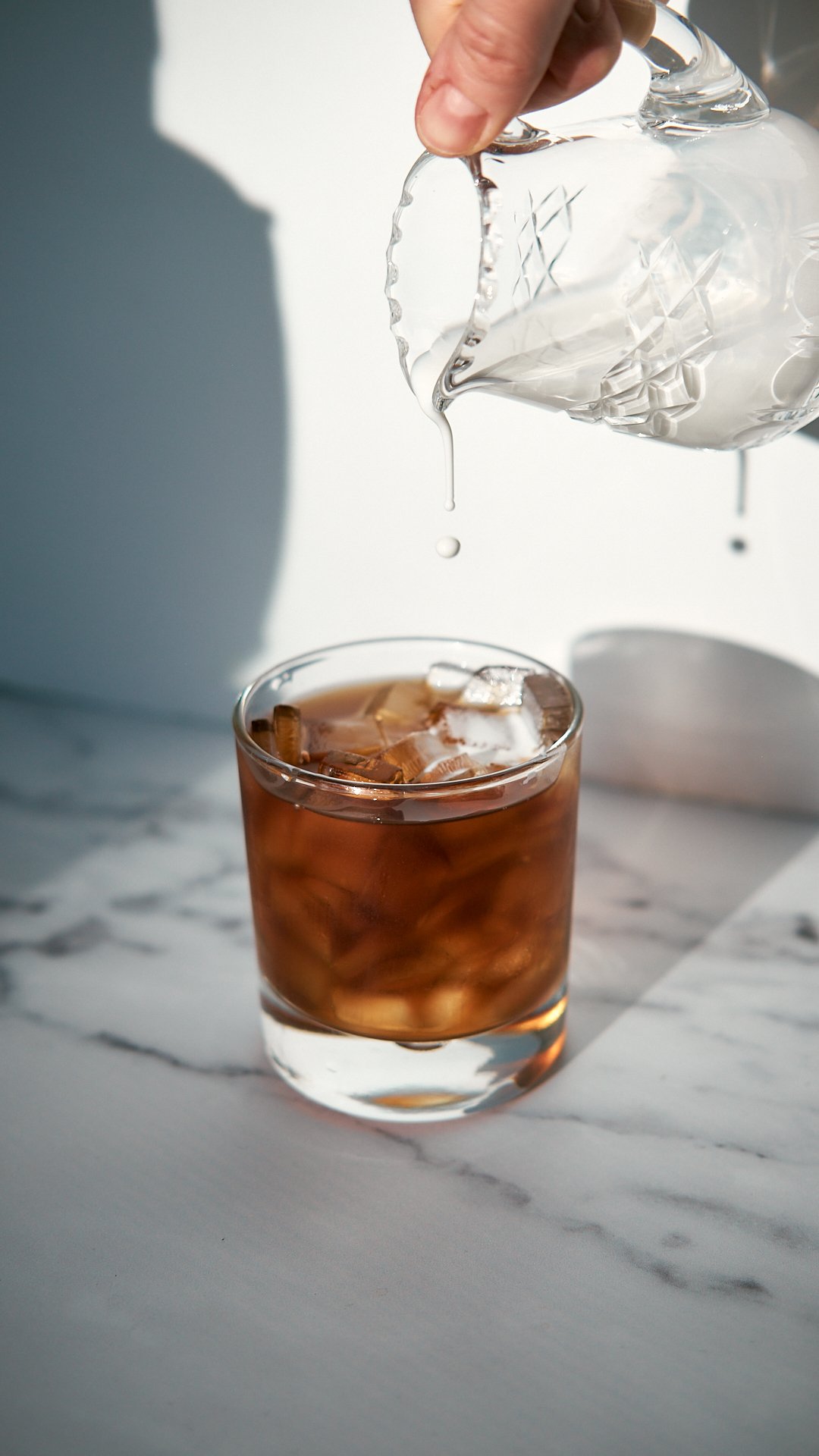 A hand pouring clear liquid into a glass of iced tea on a marble surface.