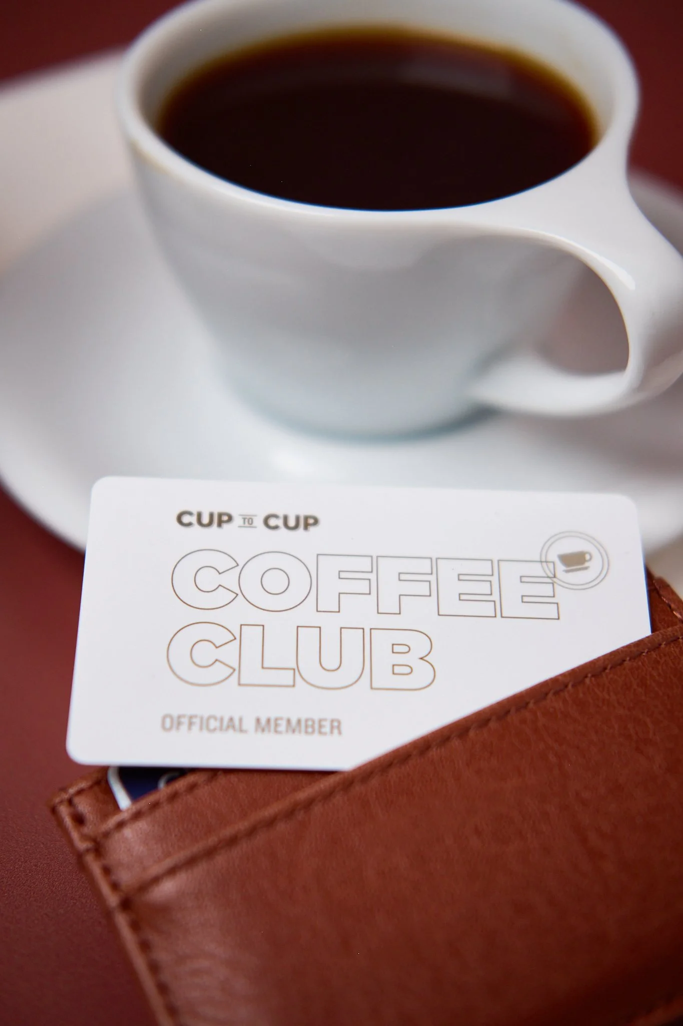 A white coffee cup filled with black coffee on a saucer, placed behind a coffee club membership card that reads 'CUP TO CUP COFFEE CLUB OFFICIAL MEMBER' and has a small coffee cup icon on the right, with a brown leather wallet partially visible under