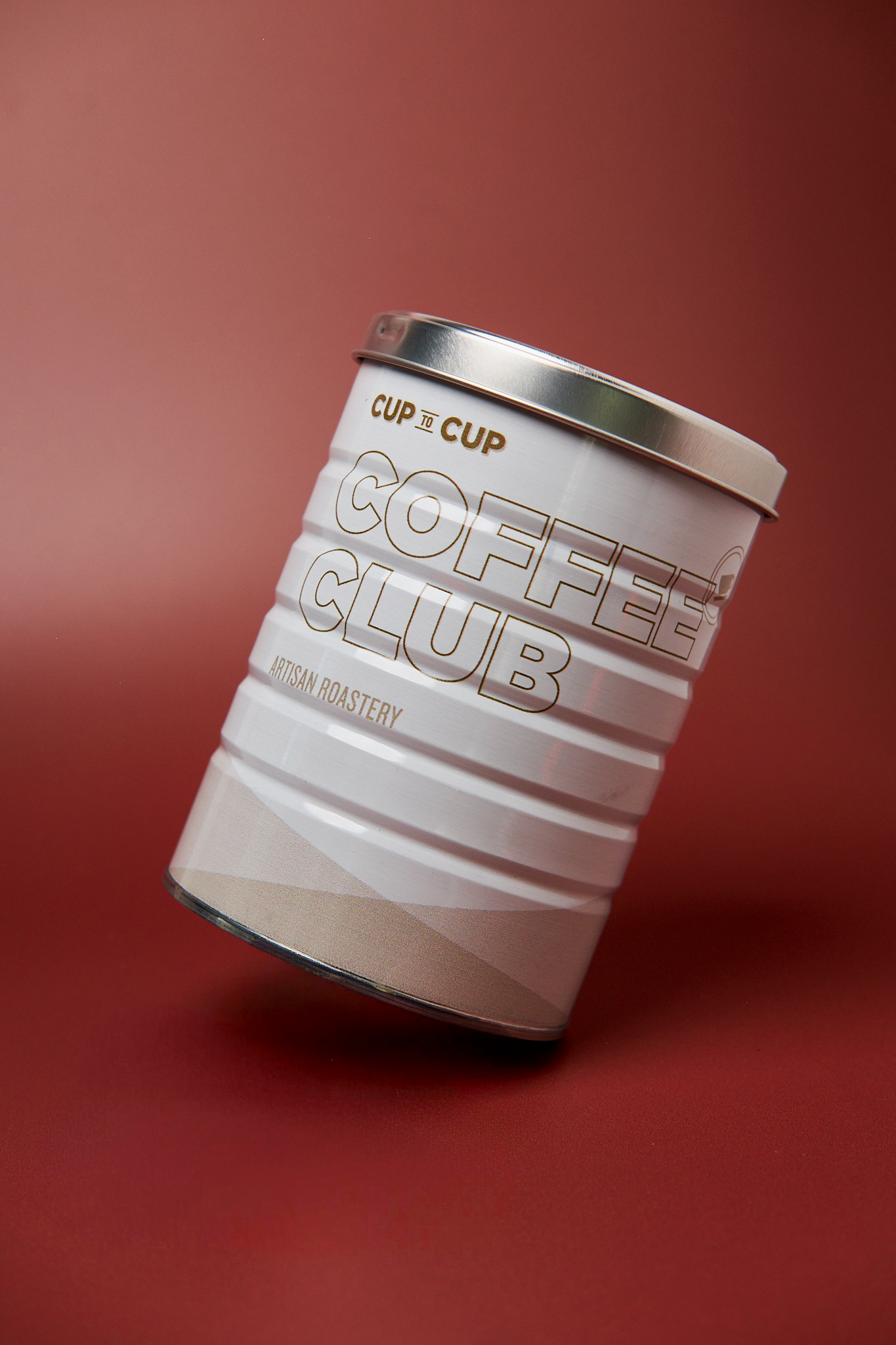 A white metal cup with the words 'Coffee Cup Club' and 'Artisan Roastery' written on it, set against a red background.