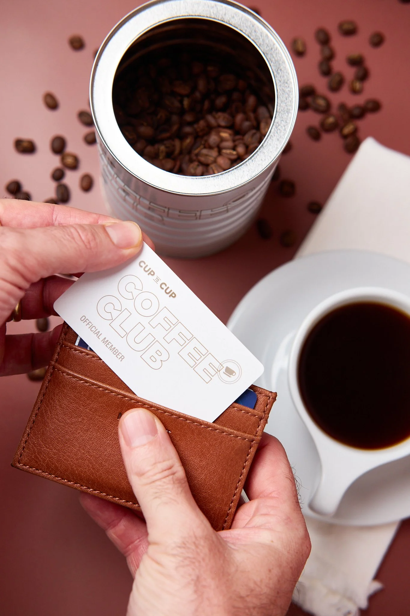 A hand holding a leather card holder with a coffee loyalty card partially pulled out, reading 'Coffee Club,' along with a small coffee cup filled with black coffee, a container of coffee beans with some spilled on the pink surface, and a white cup of