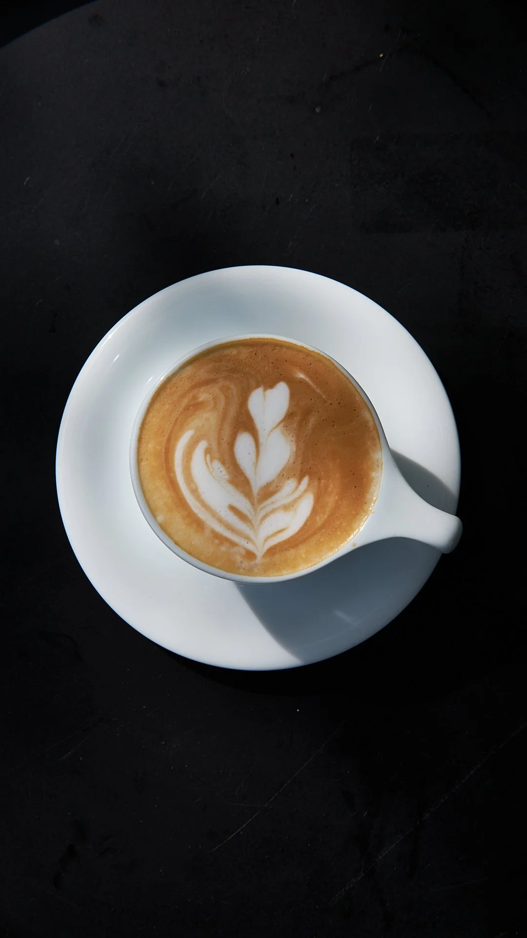 A white coffee cup with a latte art design of a feather or tulip, sitting on a matching white saucer on a dark surface.