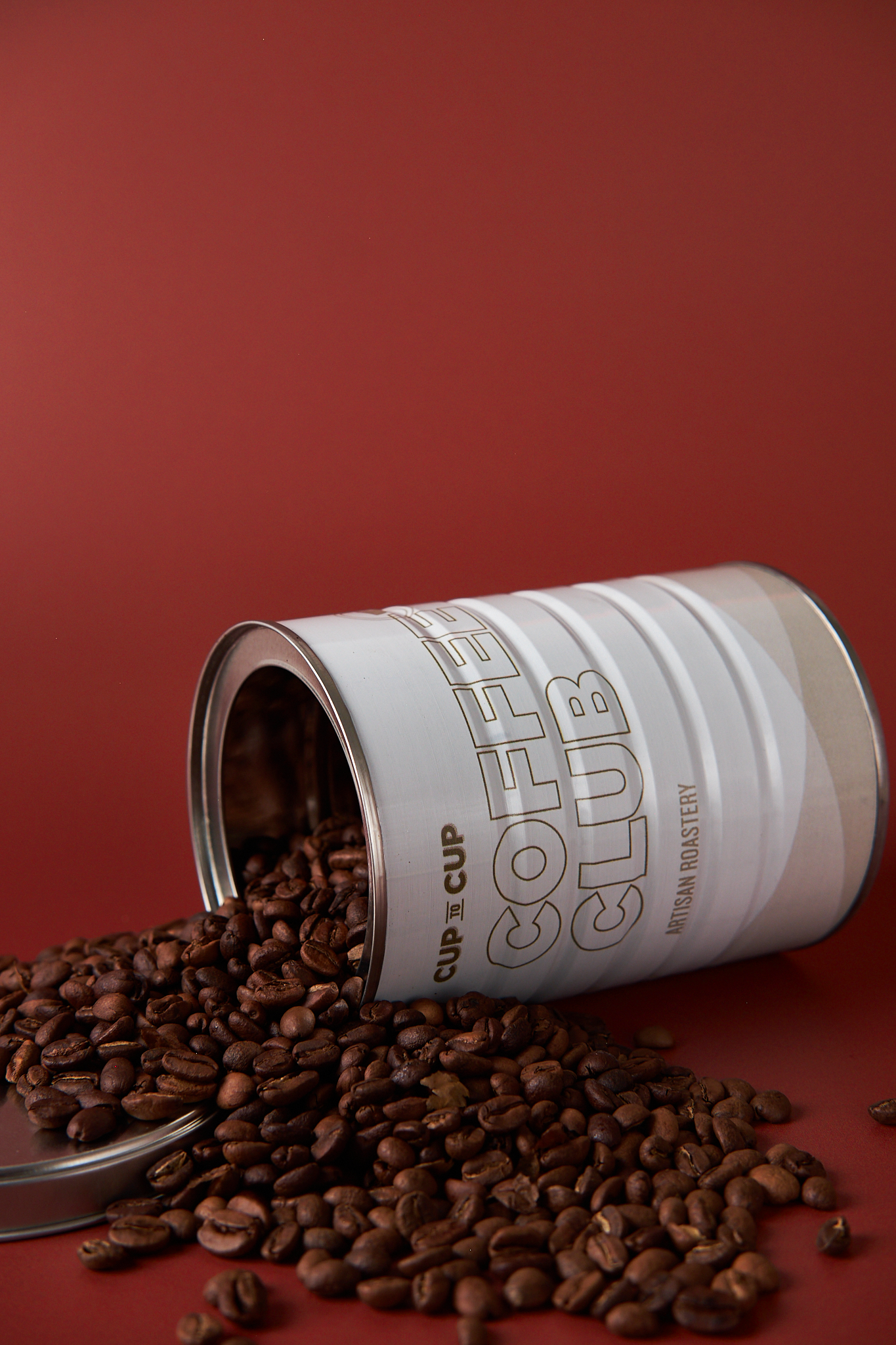 A metal can labeled 'COFFEE CLUB' with ground coffee spilling out onto a red surface and background.