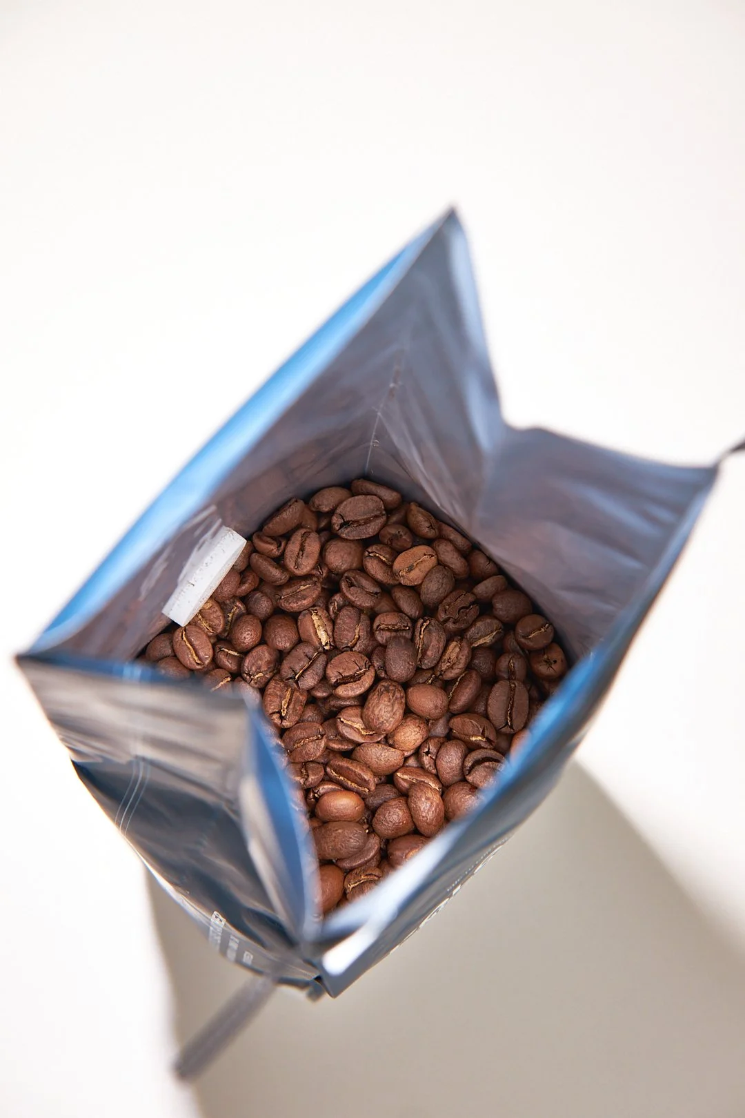 Top view of an open silver bag containing roasted coffee beans over a white surface.