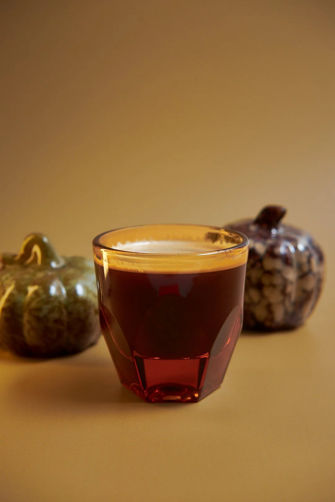 A glass filled with a dark beverage, placed on a flat surface with two decorative pumpkins in the background.