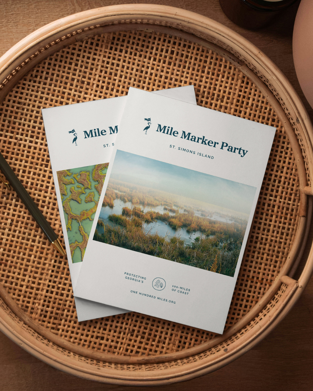 Flyers for the Mile Marker Party on St. Simons Island, featuring a landscape photo of a marsh with water and grass, placed in a basket with a pen nearby.