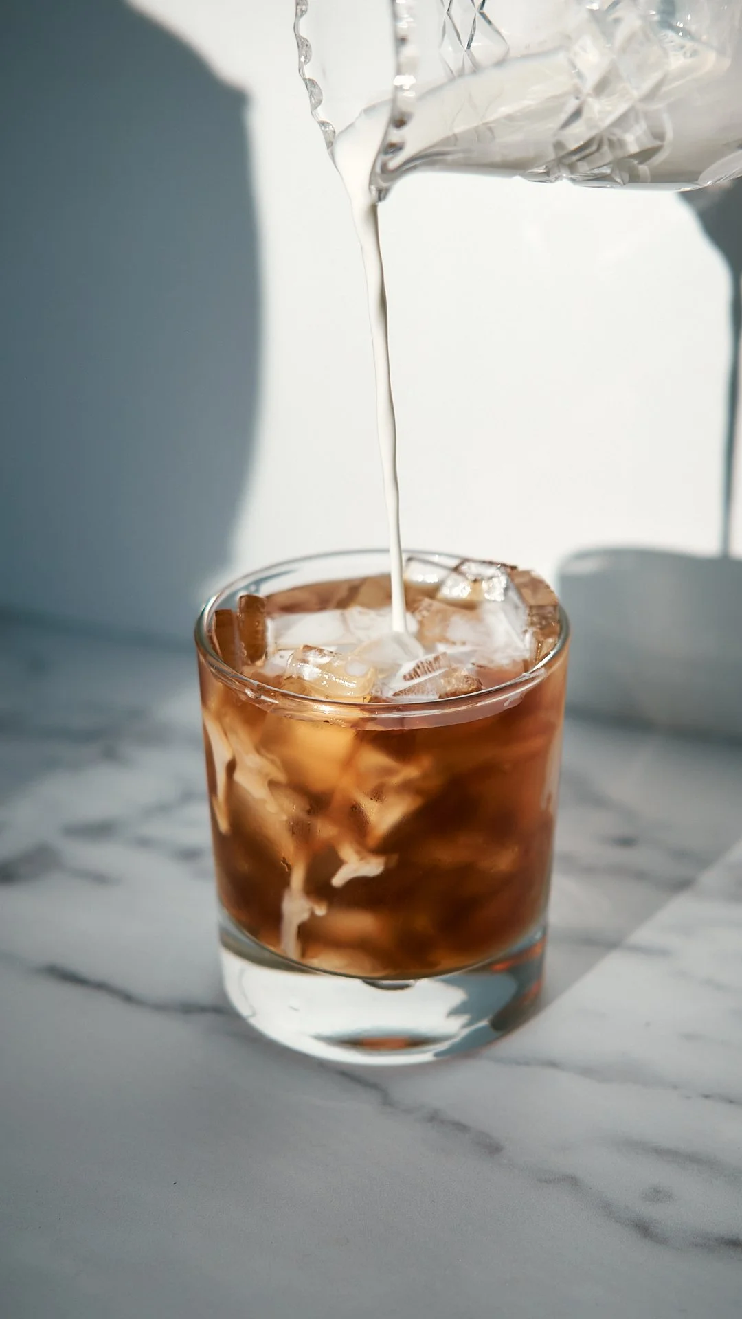 Pouring milk into a glass of iced coffee on a marble surface with a white background.