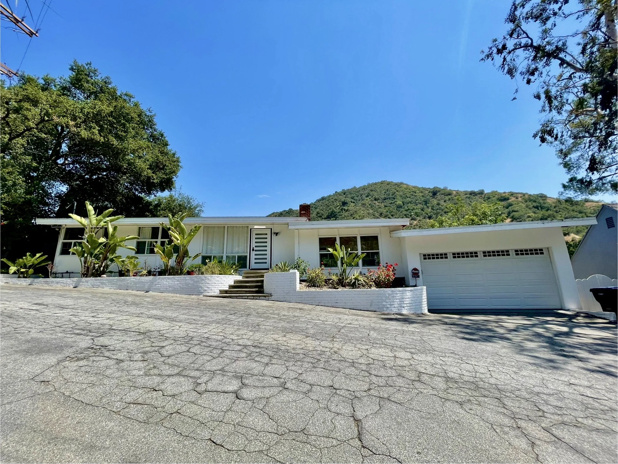 Front view of a white, mid-century modern house with a garden, driveway, and mountain in the background under a clear blue sky.
