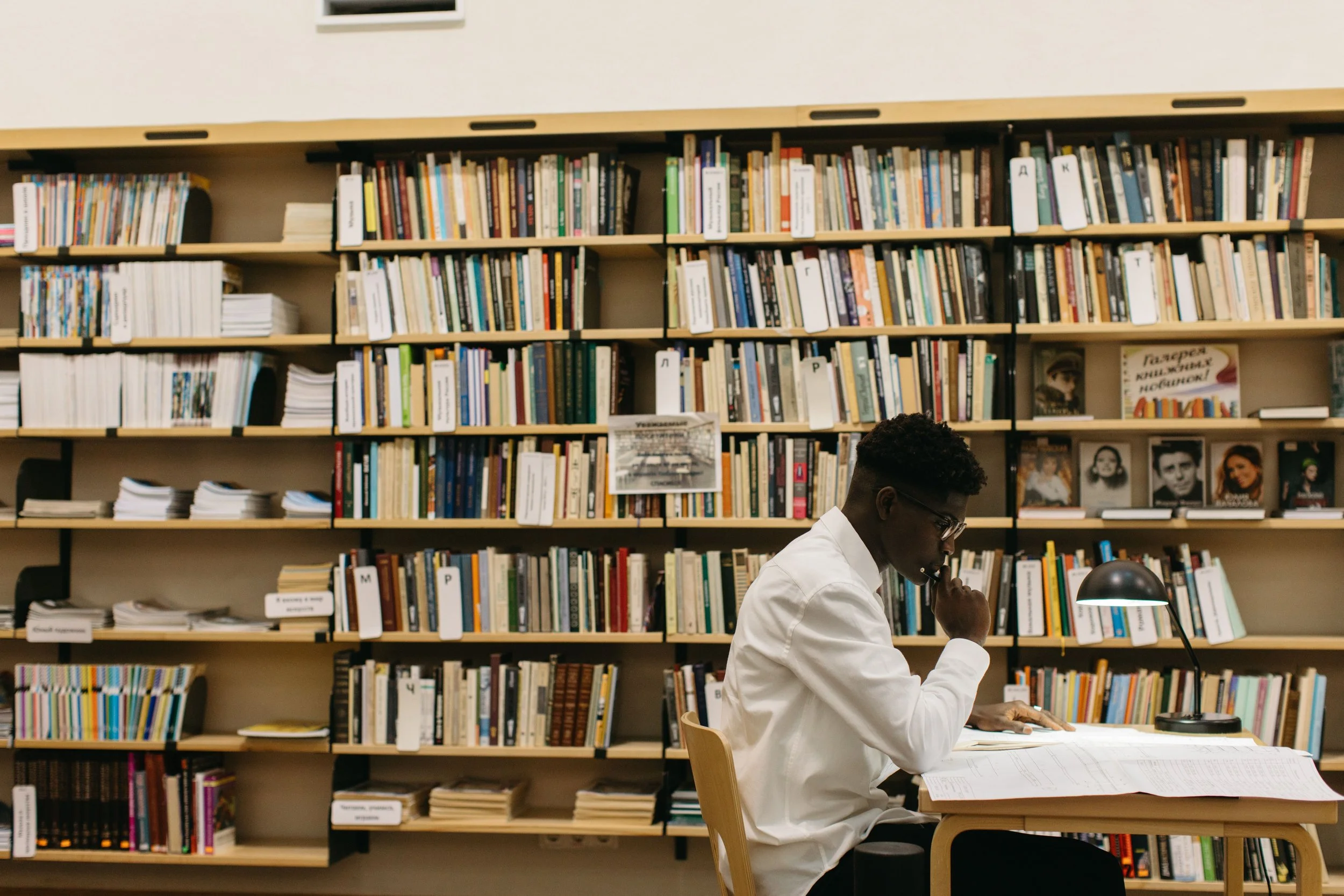 A college student in a white shirt studies at a desk lamp in a library, with tall shelves of books filling the wall behind him.