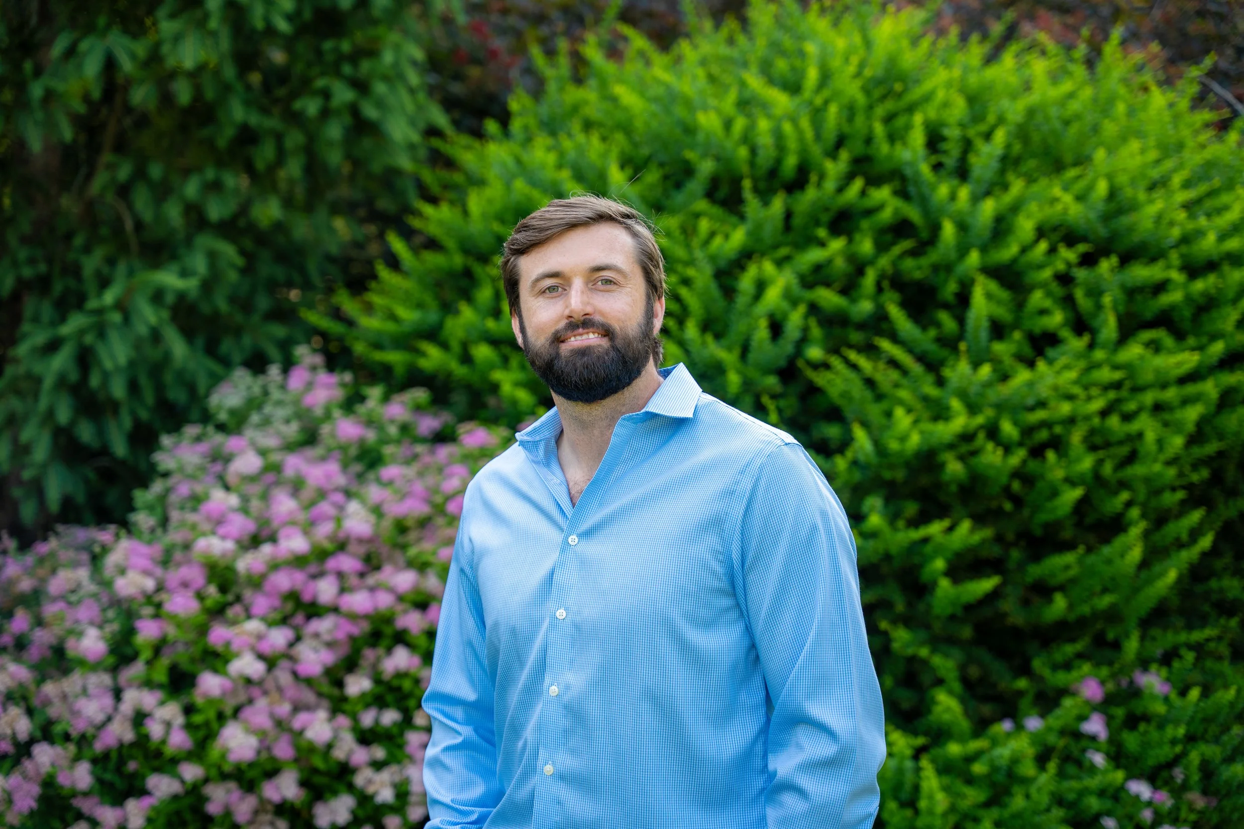 Outdoor portrait of a smiling man in a blue shirt beside flowering garden hedges