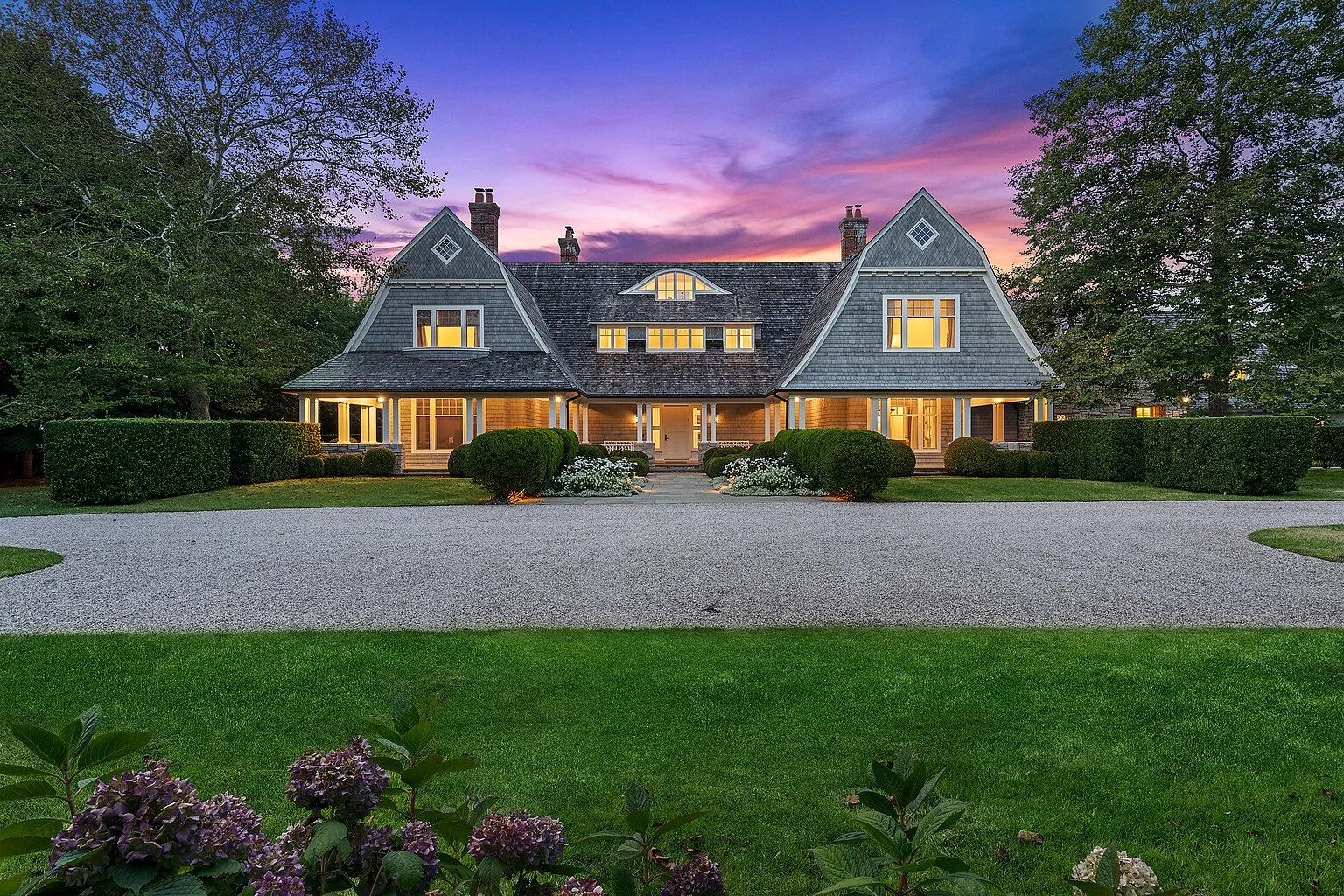 Shingle-style Hamptons estate at twilight with lit windows and a circular gravel driveway