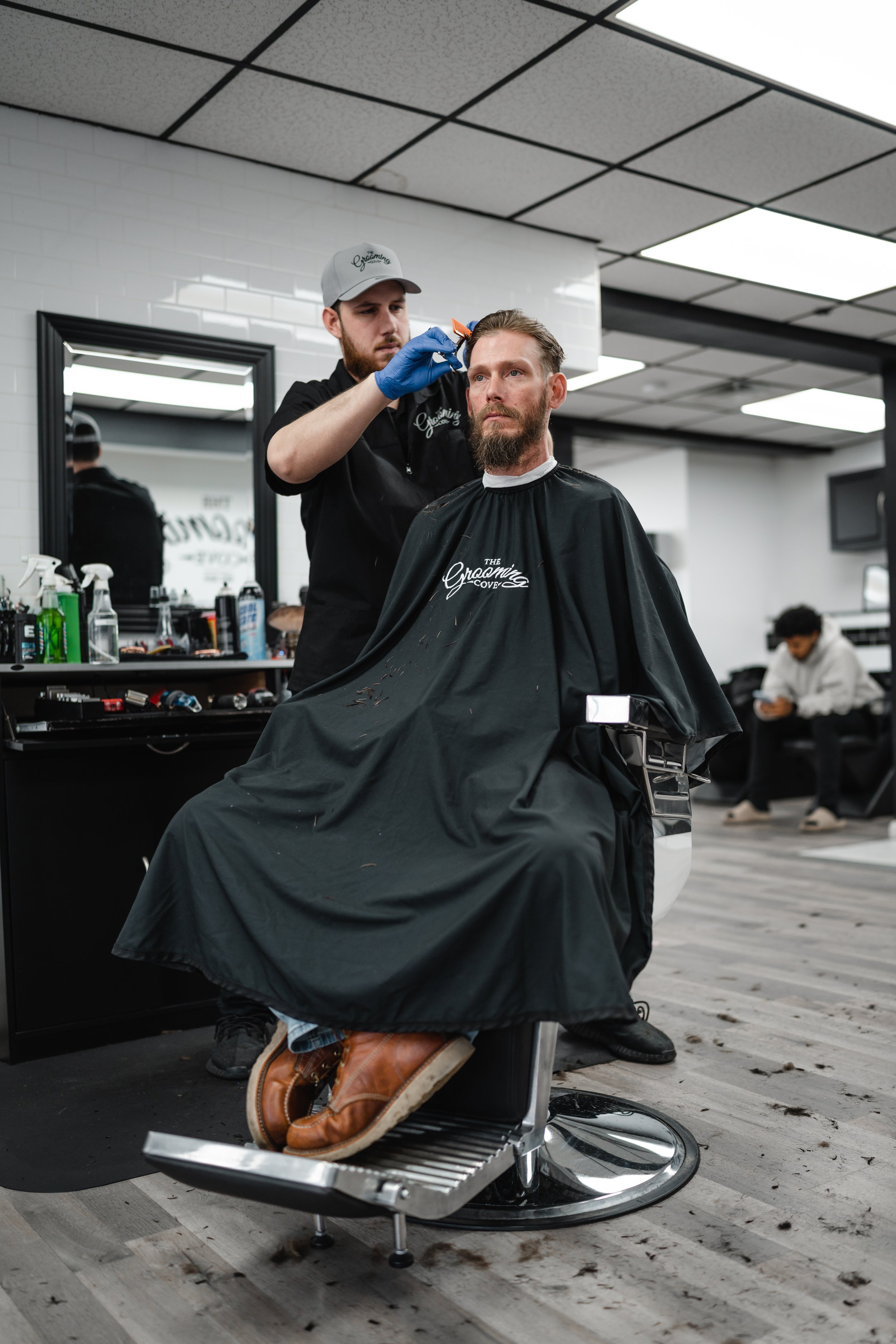 Barber cutting a man's hair in a barbershop, with hair clippings on the floor and barber supplies on a counter nearby.