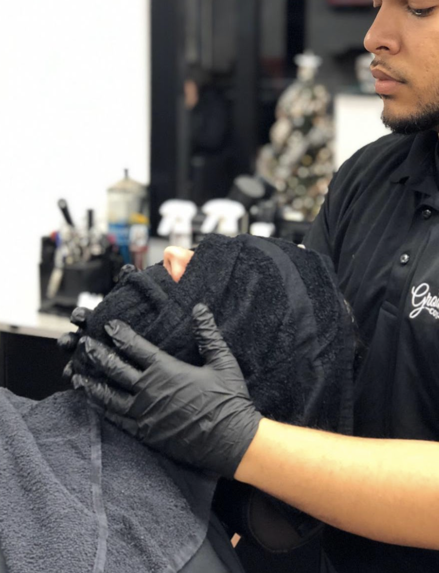 Barber holding a client's head wrapped in a towel during a haircut or grooming at a barbershop.