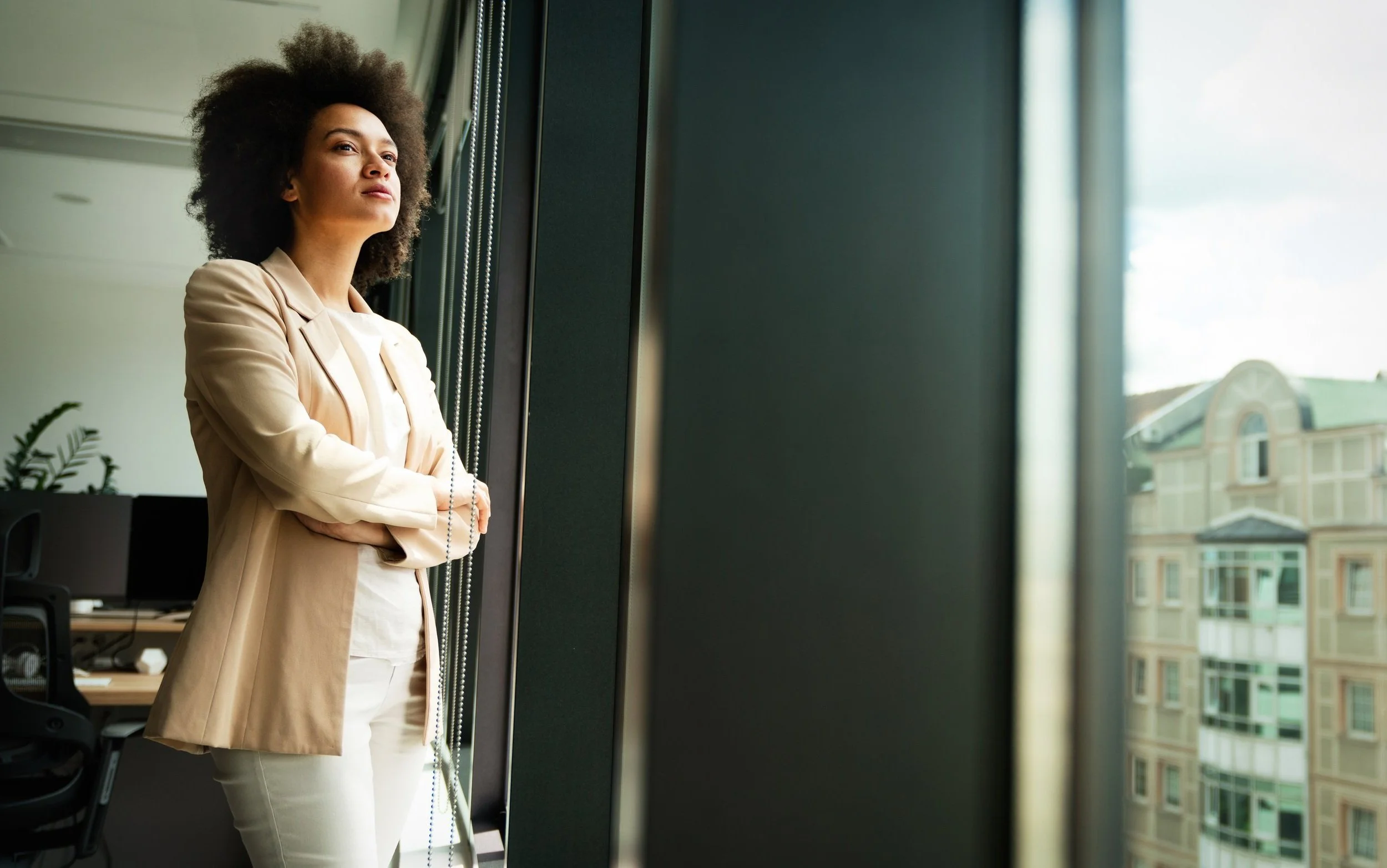 African American woman in creme suit, with arms crossed looking pensively out office window