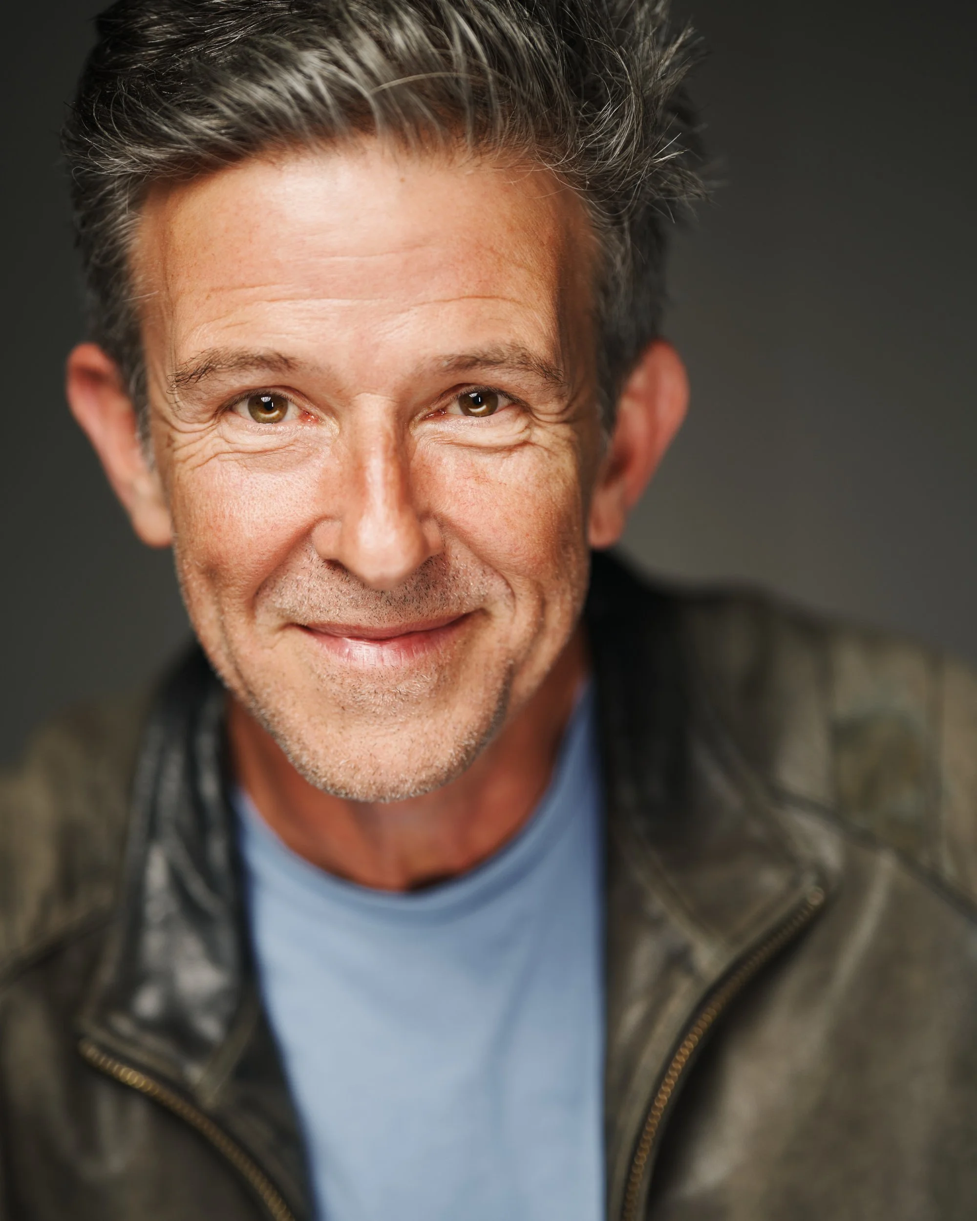 Close-up of a smiling middle-aged man with gray hair, wearing a gray zip-up hoodie and a blue shirt underneath, against a dark background.