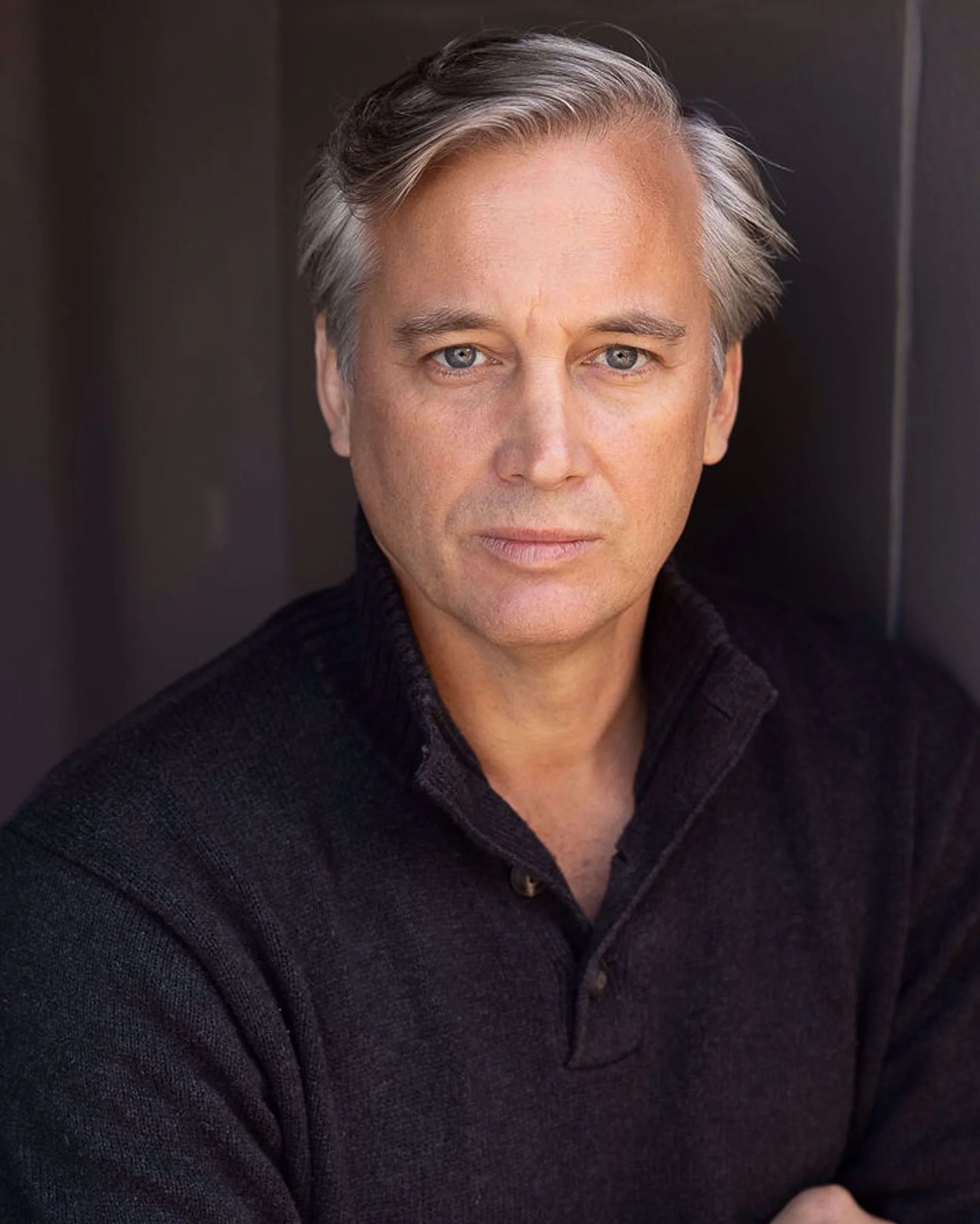 Close-up of a middle-aged man with silver hair, blue eyes, and light skin, wearing a dark textured sweater, looking directly at the camera against a dark background.