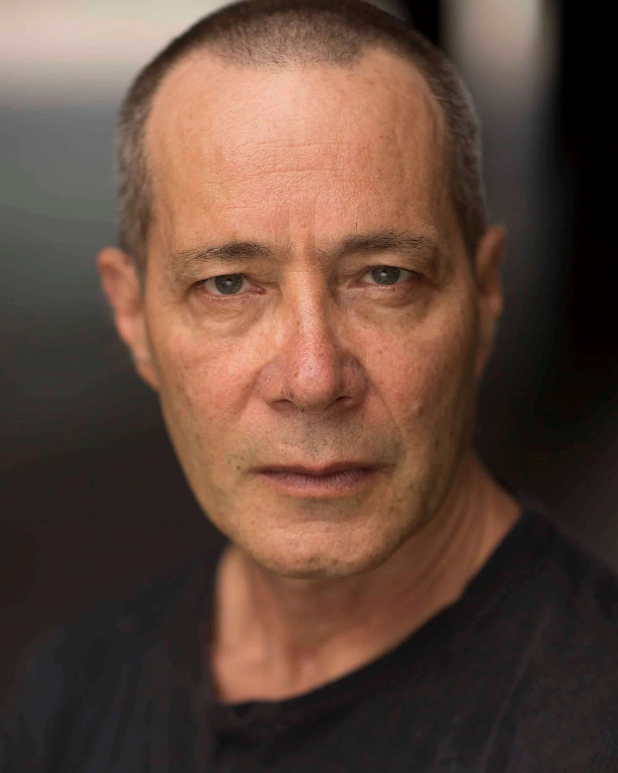 Close-up of a middle-aged man's face with a shaved head and blue eyes, wearing a black shirt, looking directly at the camera.