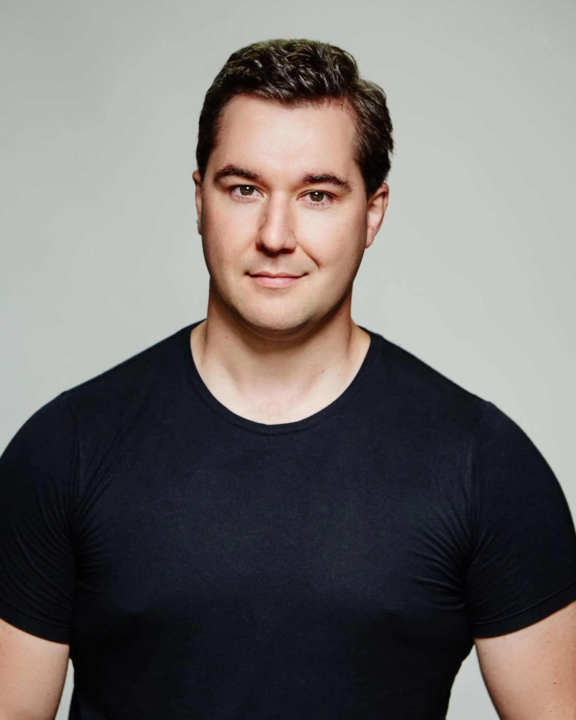 Close-up of a young man's face and upper torso, with brown hair, wearing a black t-shirt, against a plain light background.