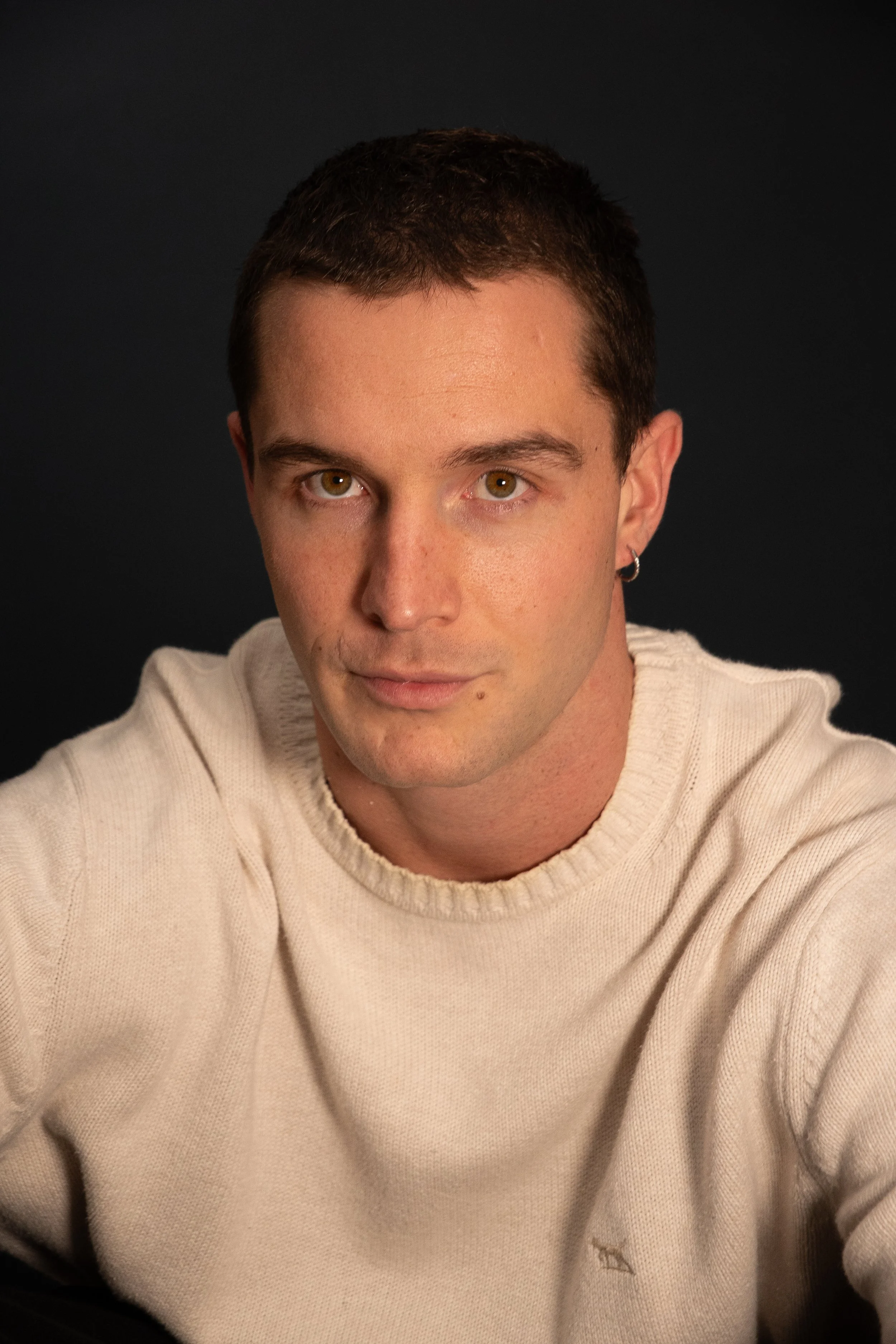 Close-up of a young man with short brown hair wearing a cream-colored sweater, looking into the camera with a neutral expression against a black background.