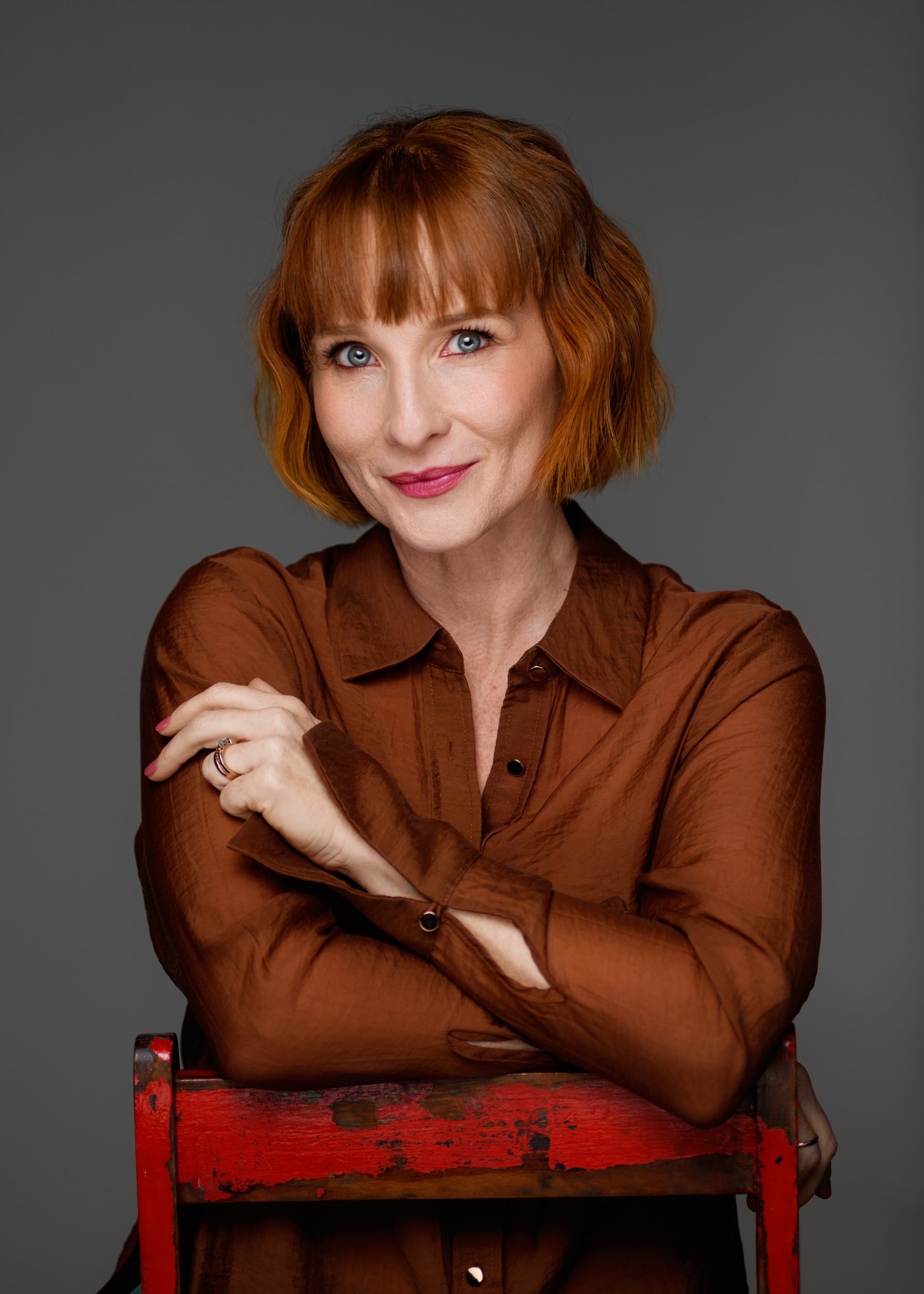 A woman with red hair and blue eyes wearing a brown shirt, smiling with arms crossed, sitting behind a red wooden chair against a gray background.