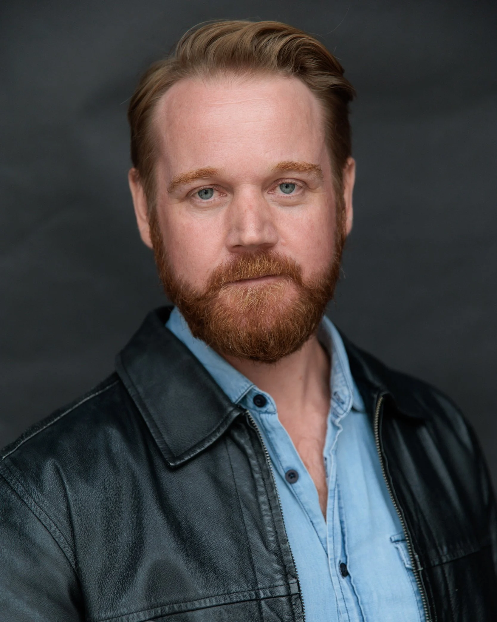 A portrait of a man with light skin, reddish hair, and a beard, wearing a blue shirt and a black leather jacket, against a dark background.